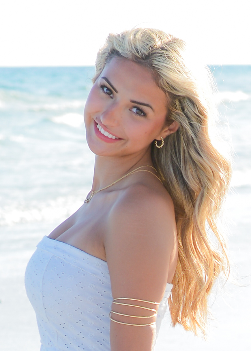 Smiling woman with long blonde hair at the beach, wearing a white strapless top and gold arm jewelry.