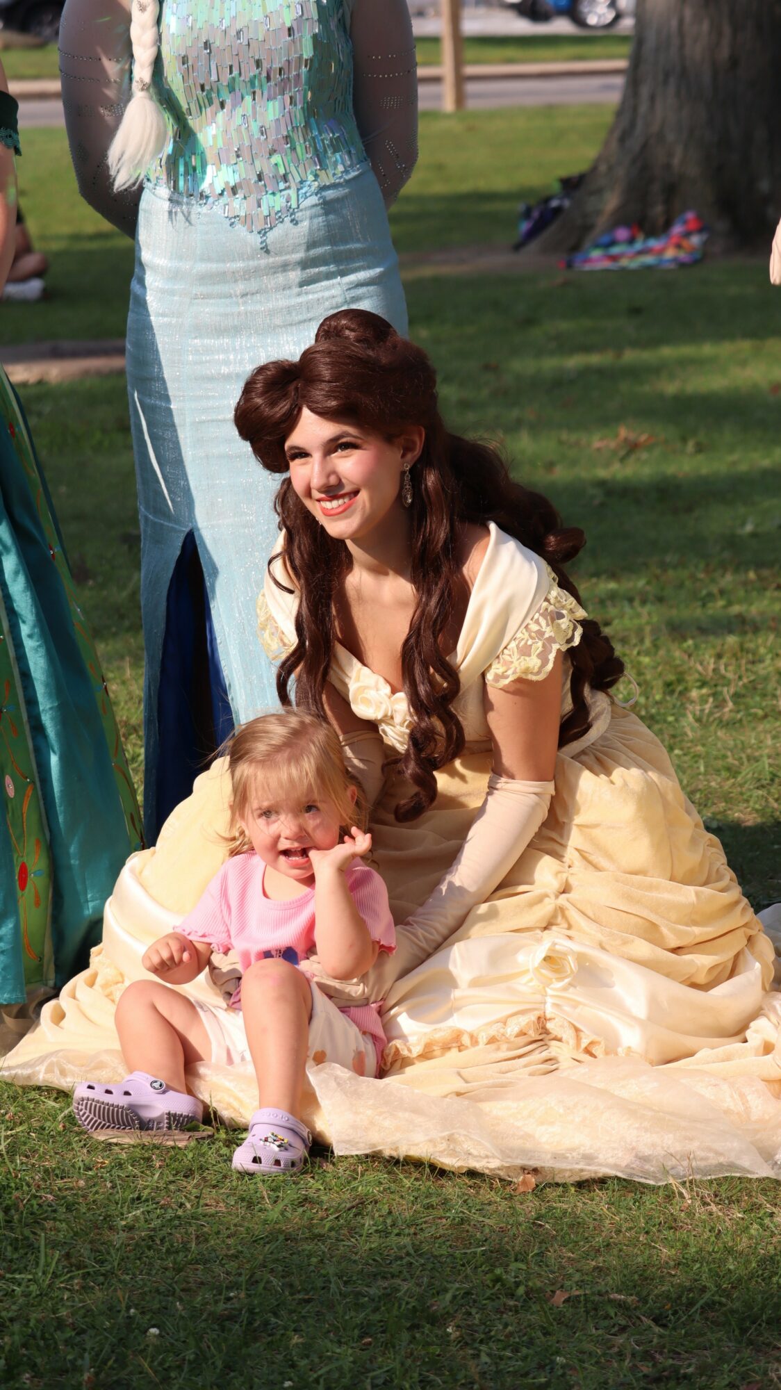 Woman in a yellow dress sitting on grass with a young girl in pink, both smiling outdoors.