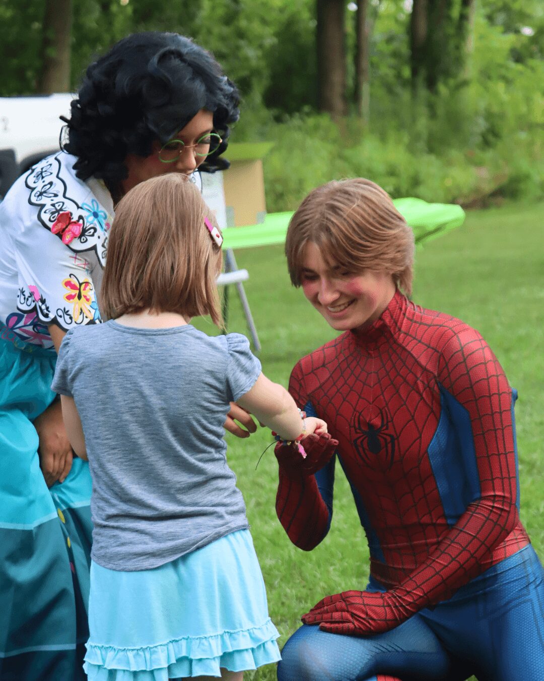 A person dressed as Spider-Man interacts with two children outdoors in a grassy area with trees, smiling and engaging.