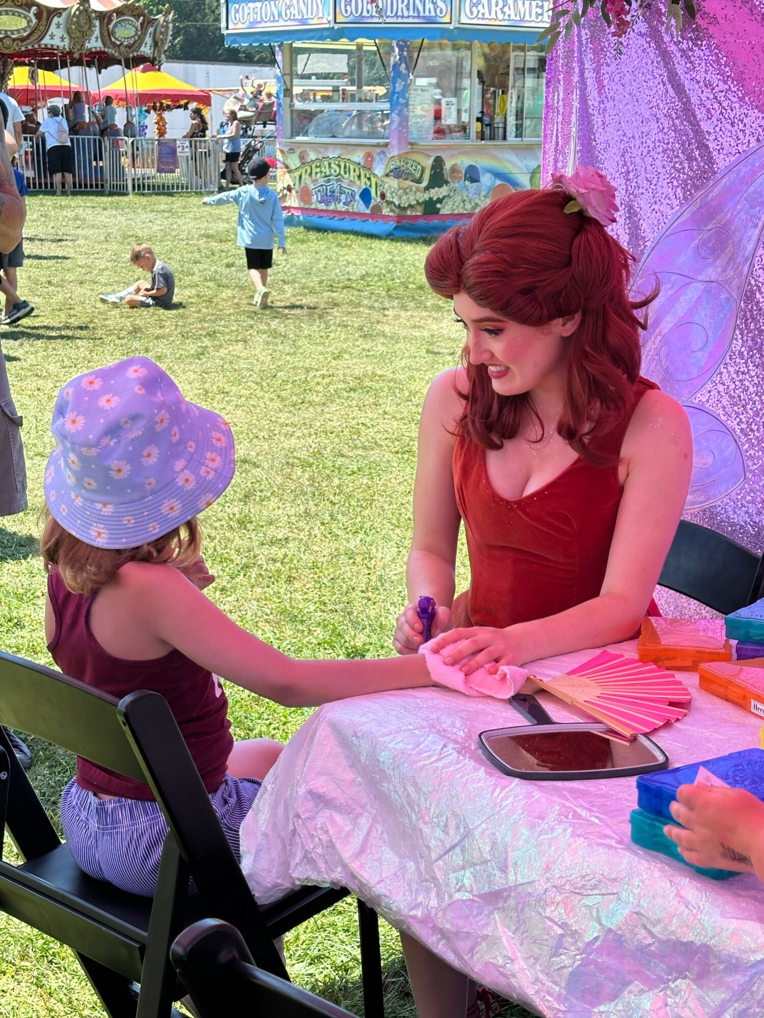 Woman with red hair and a girl with a purple hat sitting at a table outdoors, holding hands, at a fair or festival.