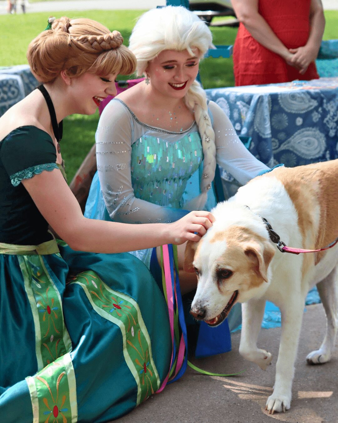 Two women dressed as princesses pet a large dog outdoors, with a table and person in red in the background.