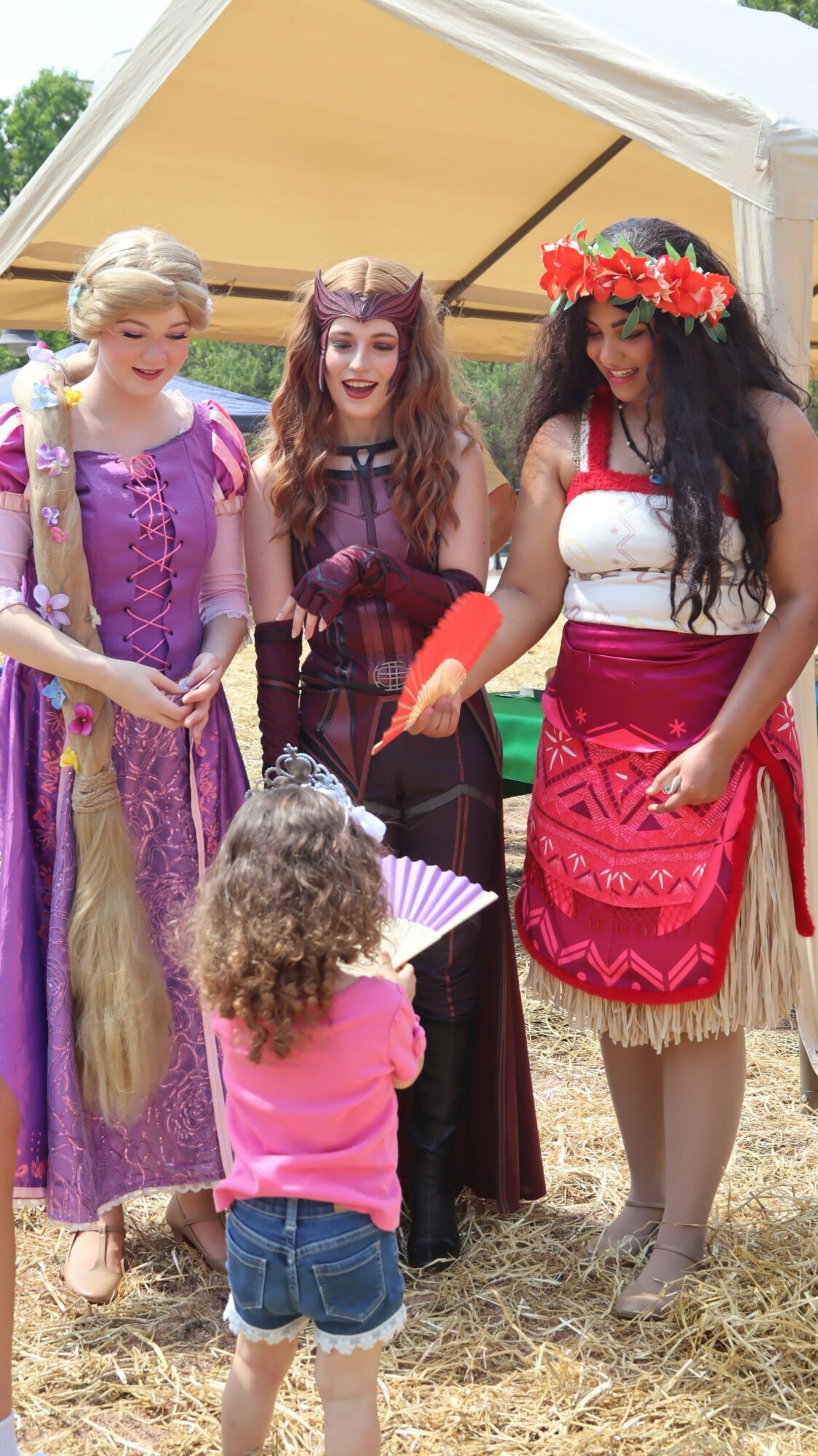 Group of women and children in colorful costumes, standing outdoors on grass, with a yellow building in the background.