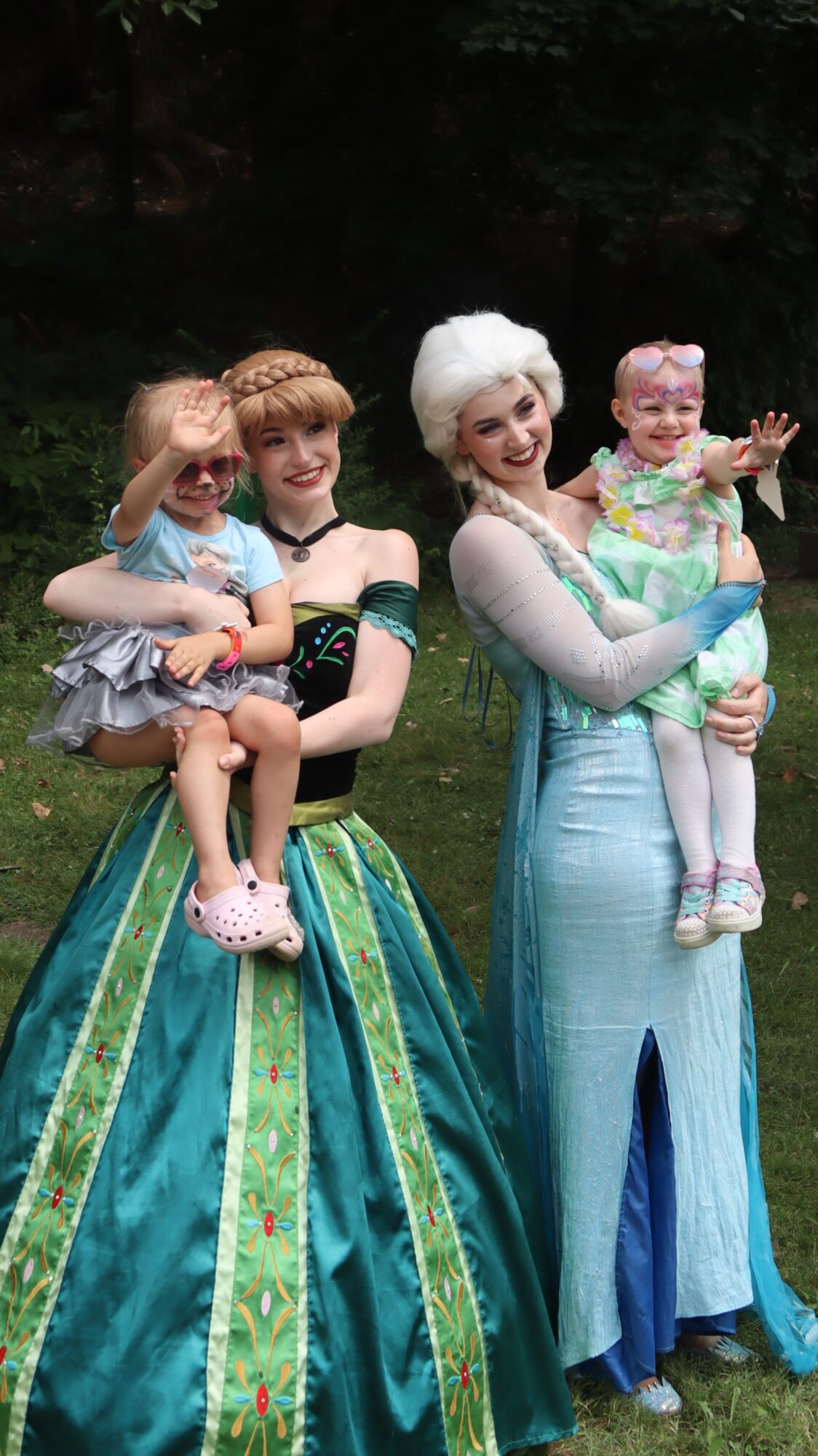 Two women dressed as princesses holding children outdoors, with trees in the background.