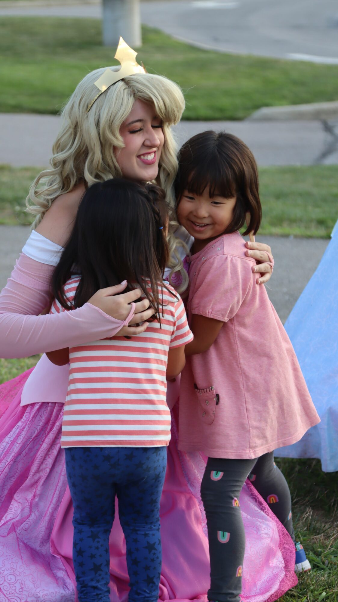 Two children and an adult outdoors, with the adult holding the children's shoulders, smiling and talking, grass and trees in background.