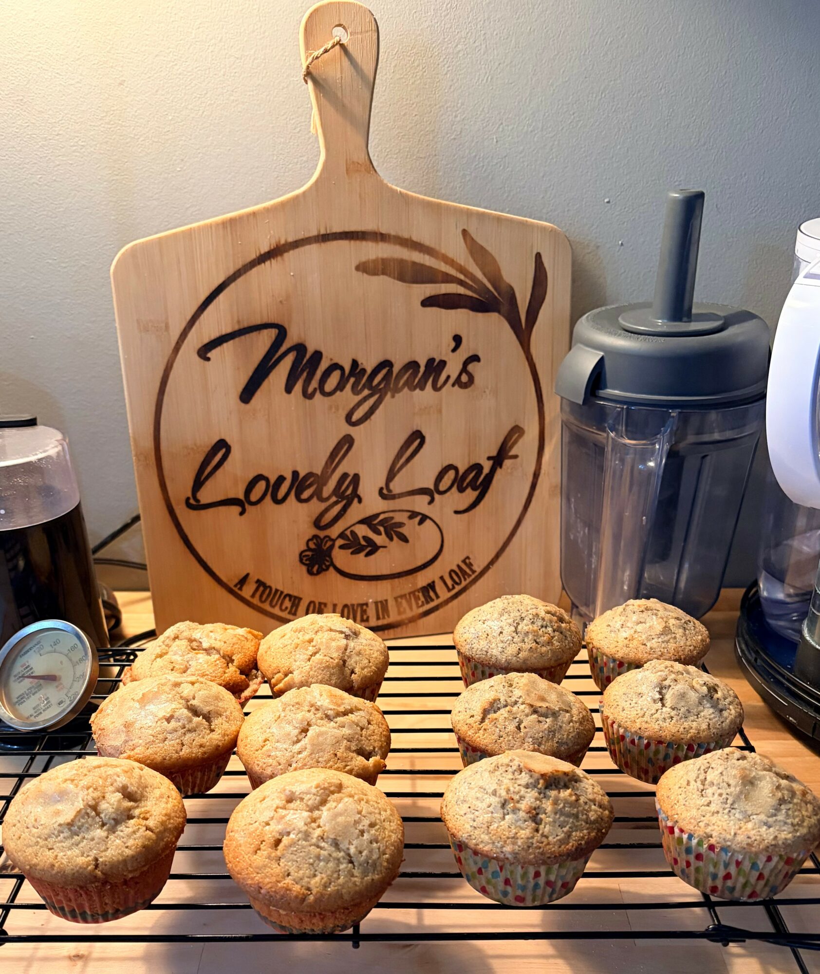 Tray of muffins in front of a wooden sign reading 'Morgan's Lovely Loaf' with a wheat illustration, on a kitchen counter.