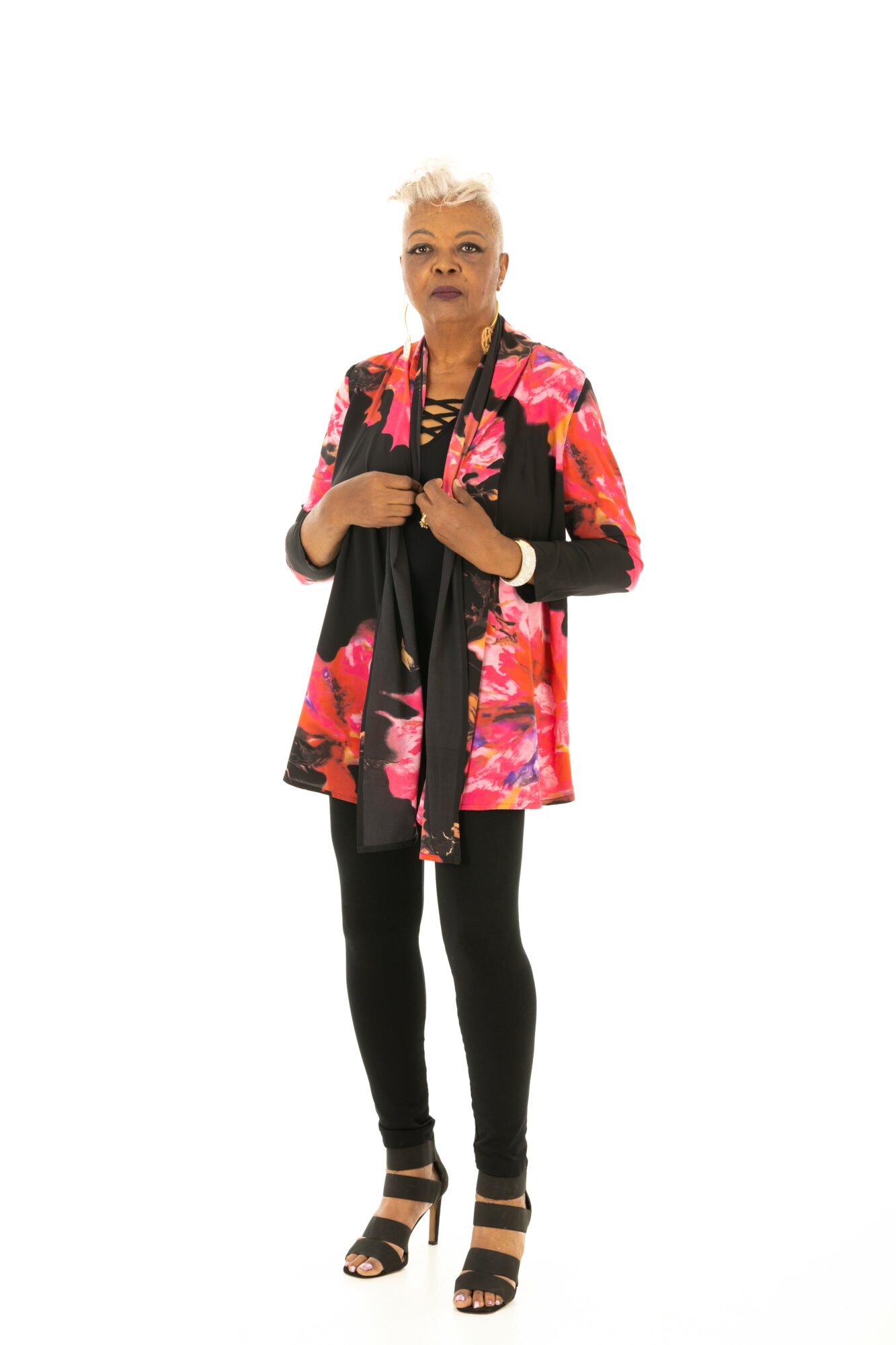 Woman with short platinum hair wearing a colorful floral jacket, black top, black leggings, and sandals, standing against a white background.
