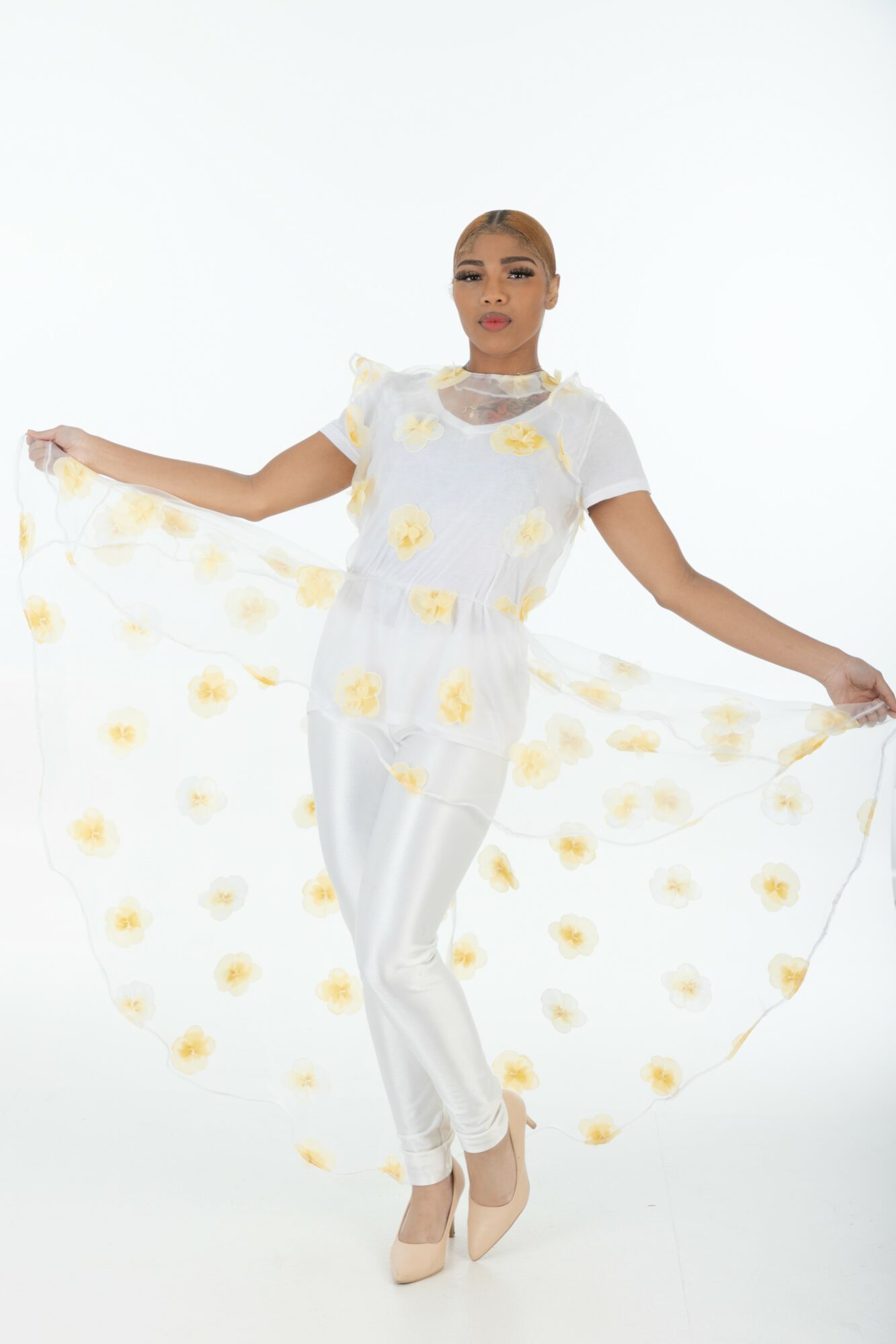 Woman in white dress holding a sheer fabric with yellow floral pattern, standing against a white background.
