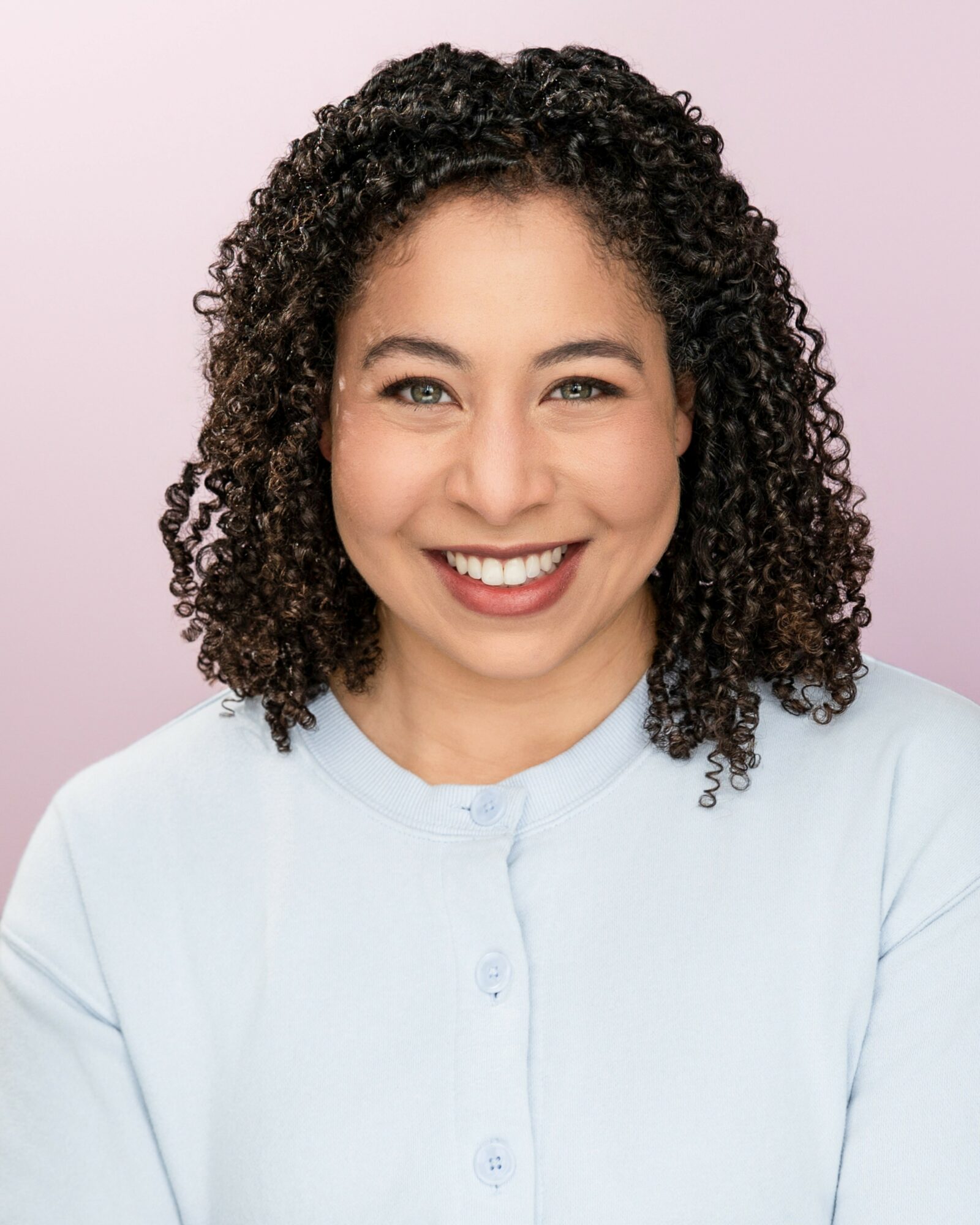 Smiling woman with curly hair wearing a light blue top, against a pink background.