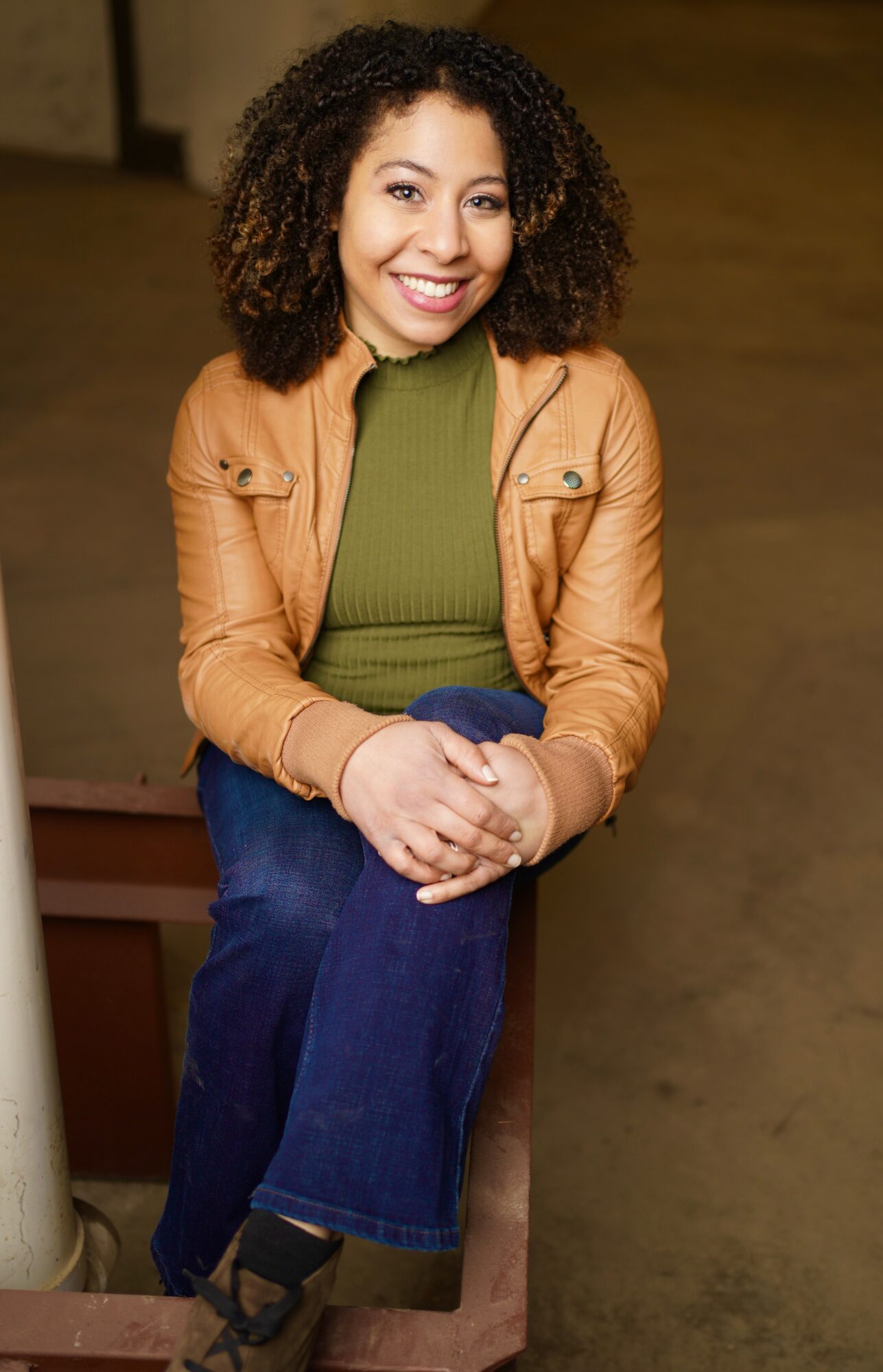 Young woman with curly hair smiling, sitting on a wooden surface, wearing a green top and tan jacket.