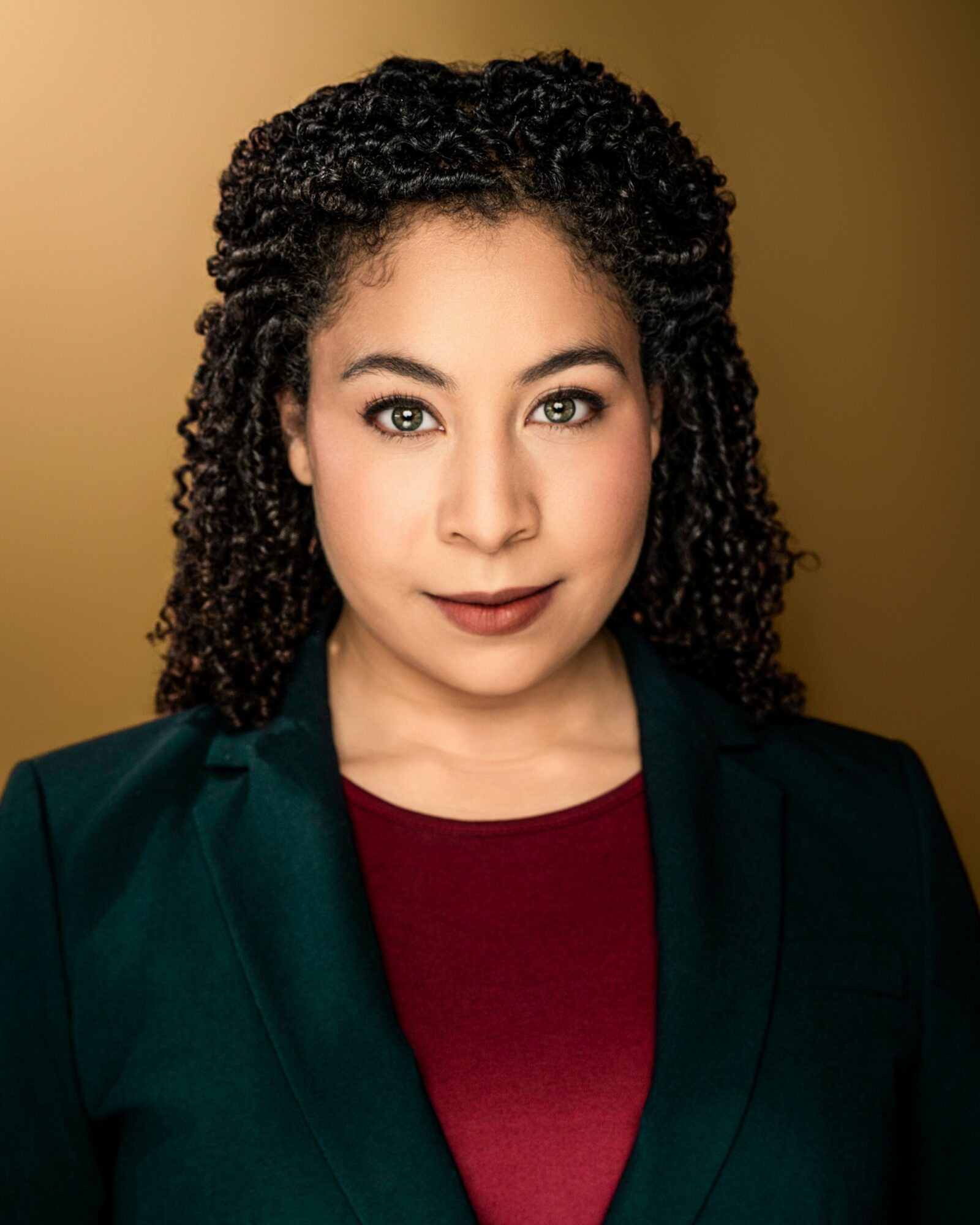 Portrait of a woman with curly hair, wearing a dark blazer and maroon top, against a warm background.