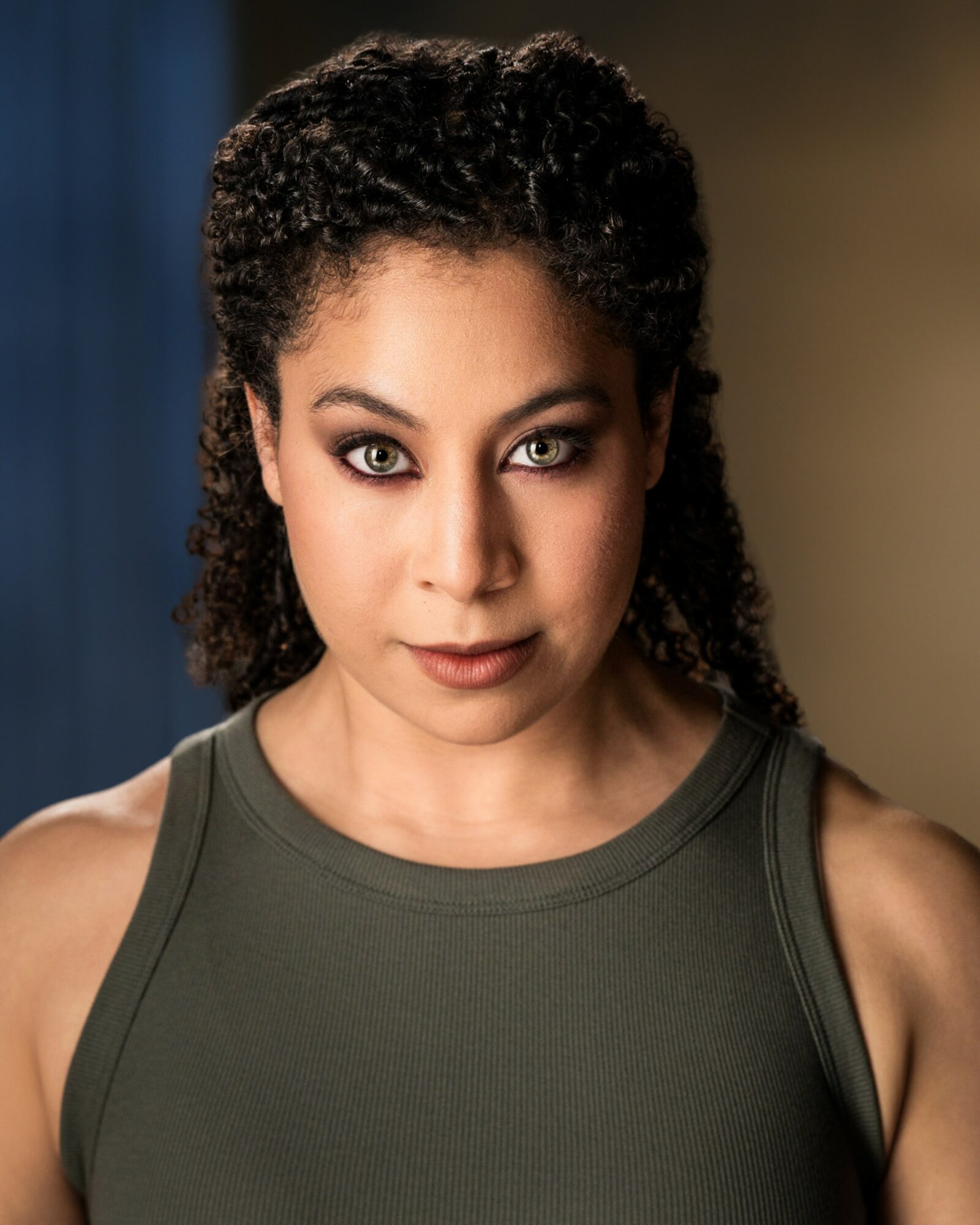 Portrait of a woman with curly hair and intense gaze, wearing a sleeveless top, against a blurred background.