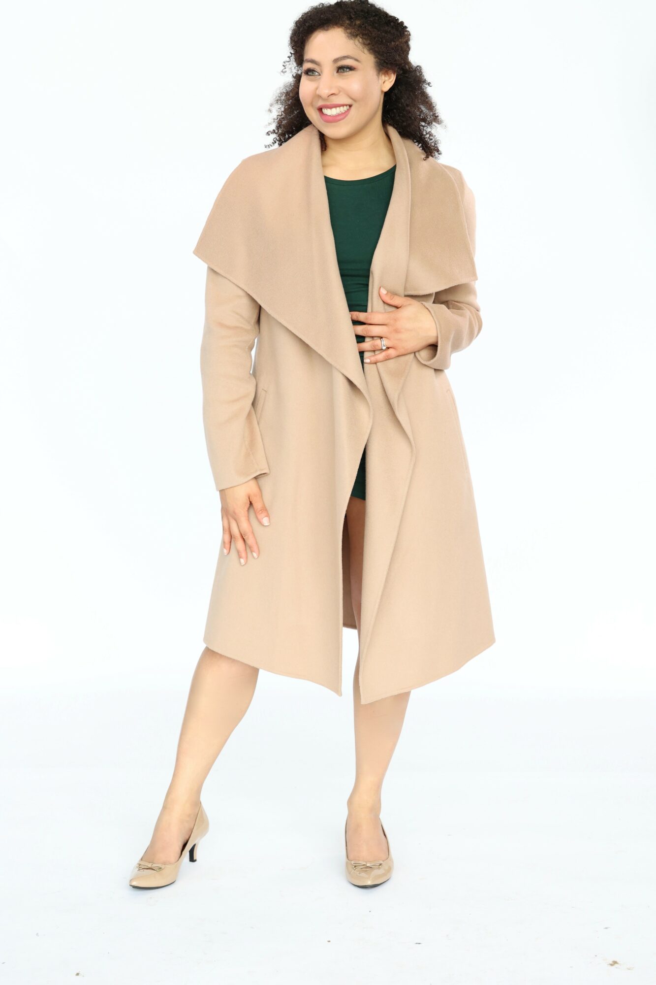 Woman with curly hair smiling, wearing a beige coat over a green top, standing with one hand on her stomach, white background.
