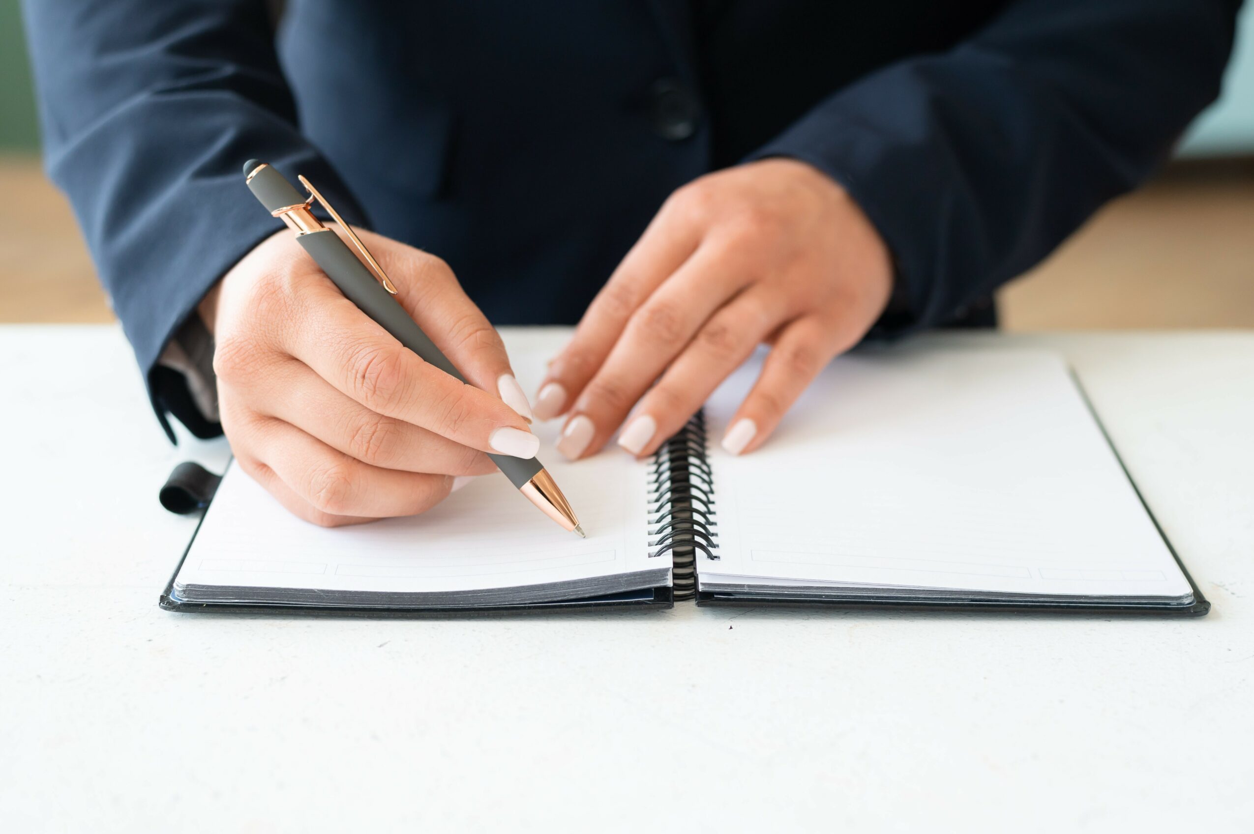 Person in a suit writes in a notebook with a pen on a white surface.