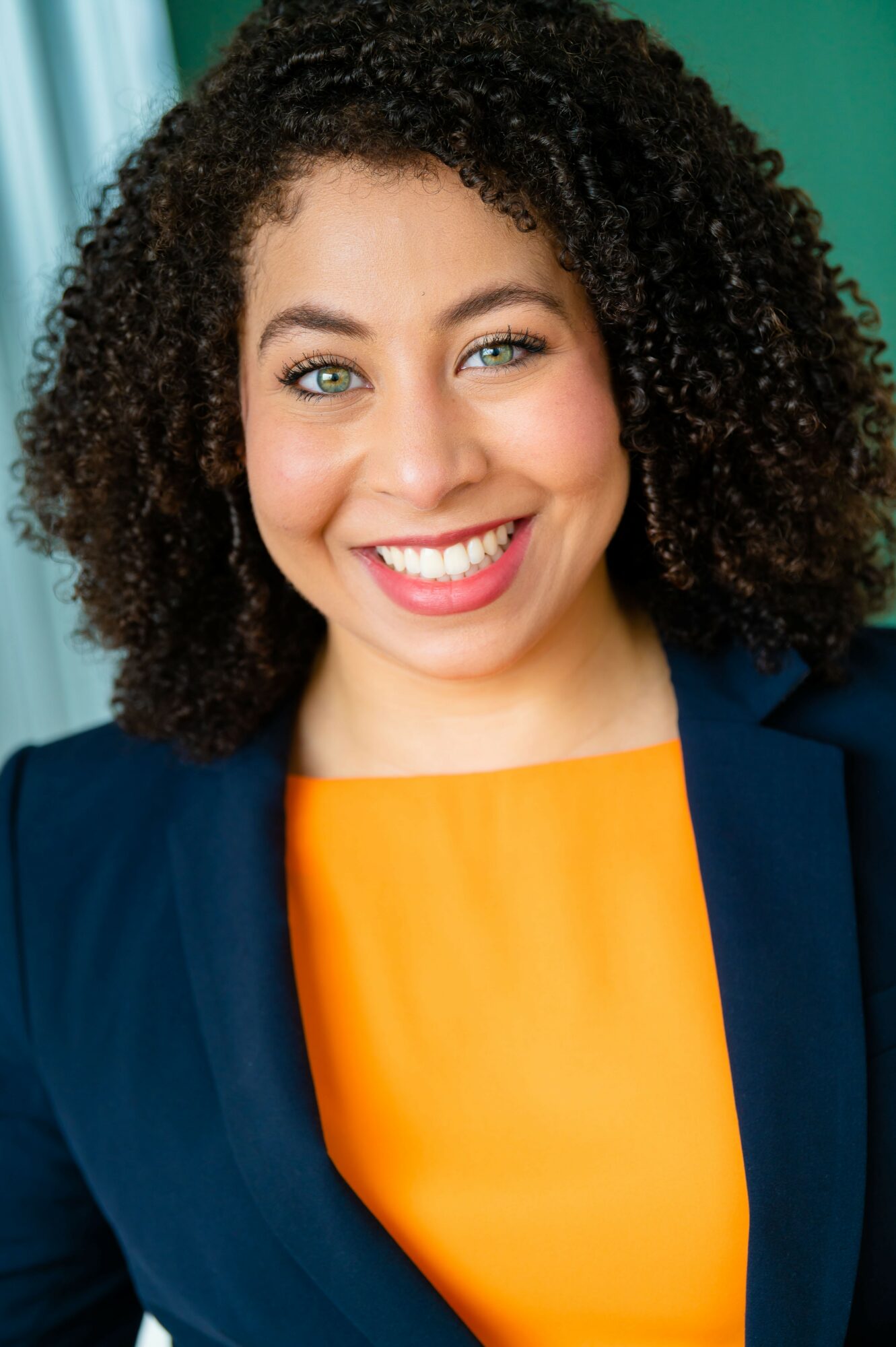Smiling woman with curly hair, wearing a navy blazer and orange top, in front of a green background.