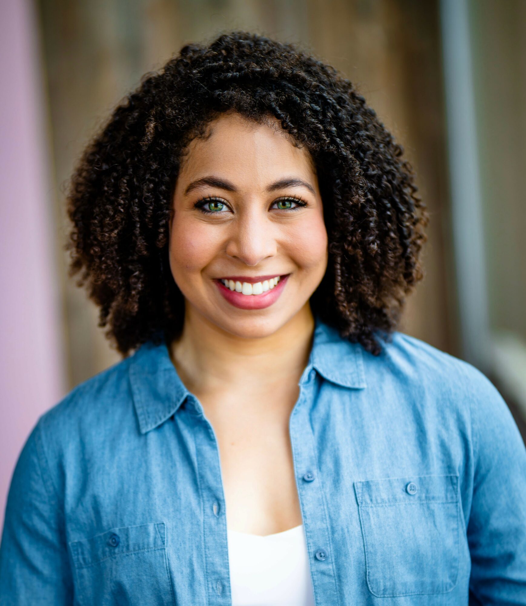 Smiling woman with curly hair wearing a blue shirt indoors, background with curtains and window.