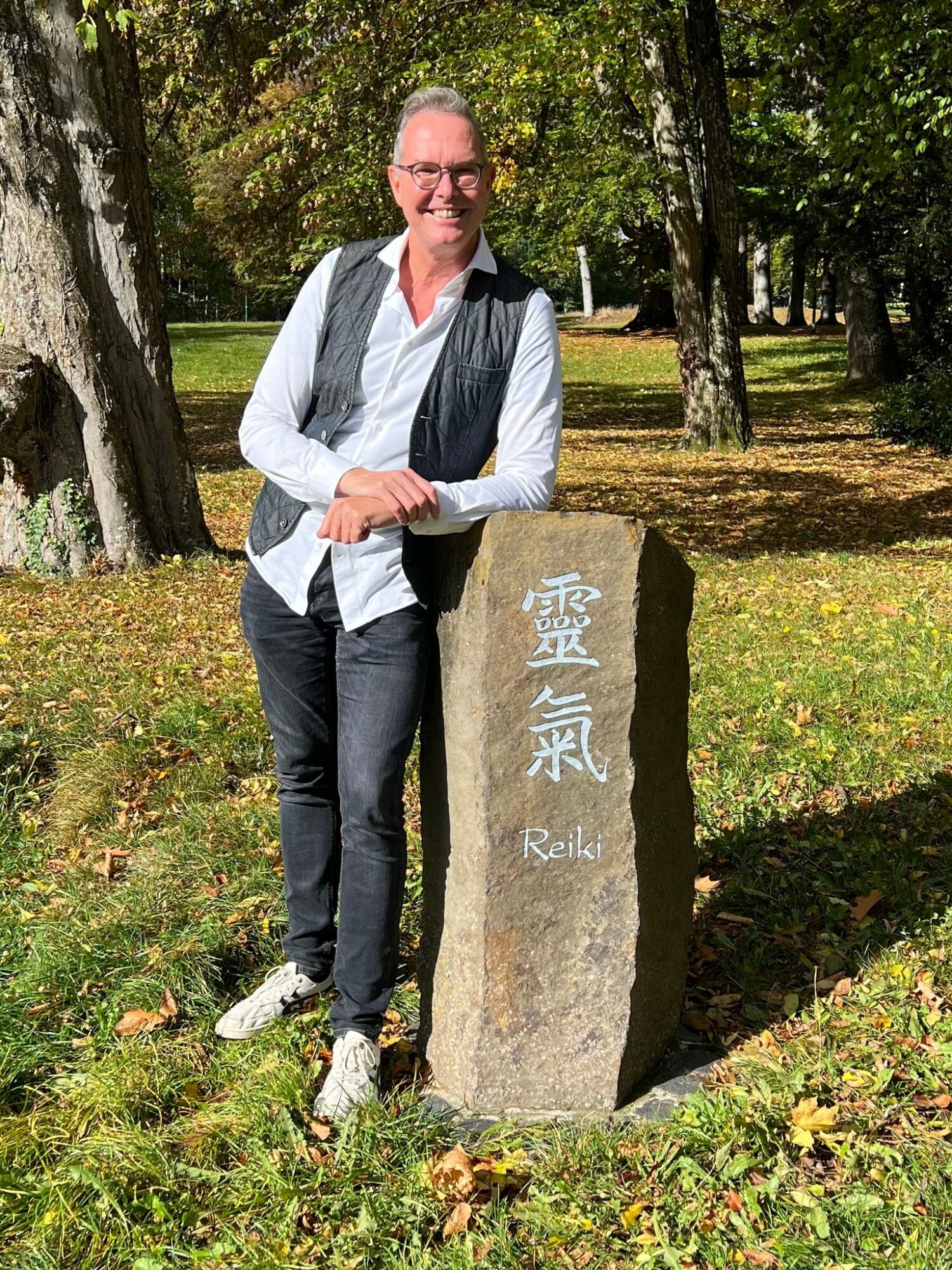 Man standing outdoors next to a stone with Japanese characters and the word 'Reiki', surrounded by trees and grass.