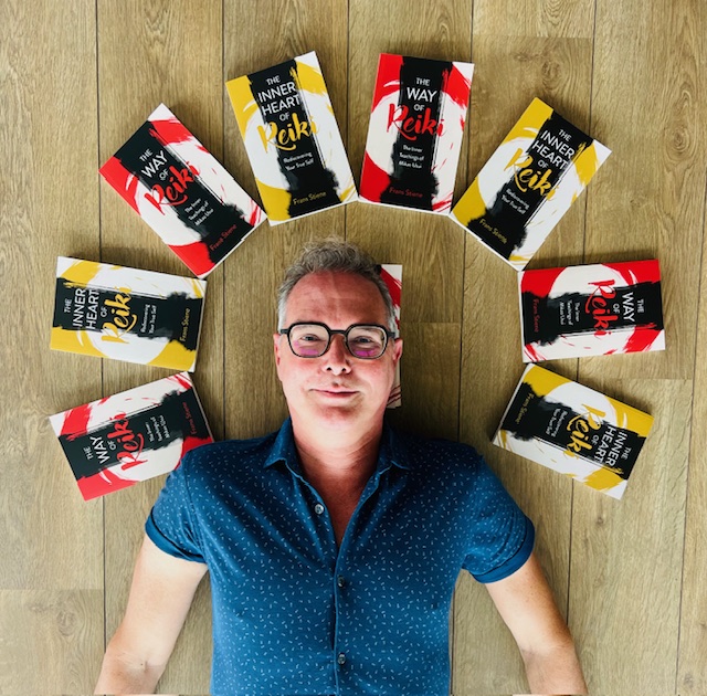 Man with glasses lying on wooden floor surrounded by books arranged in a circle, looking up at the camera.