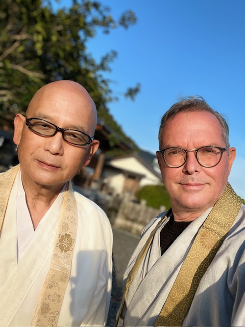 Two men wearing traditional robes and glasses outdoors, with trees and a building in the background, under a clear blue sky.