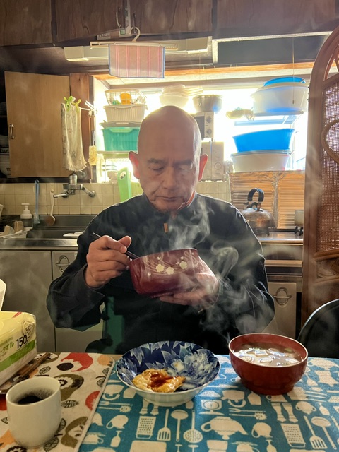 Bald man eating at a table with bowls and a cup, kitchen background with colorful containers, steam rising from food.