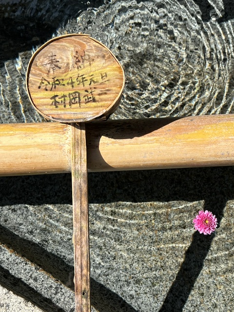 A wooden sign with Japanese characters, a pink flower, and shadows on a textured surface.