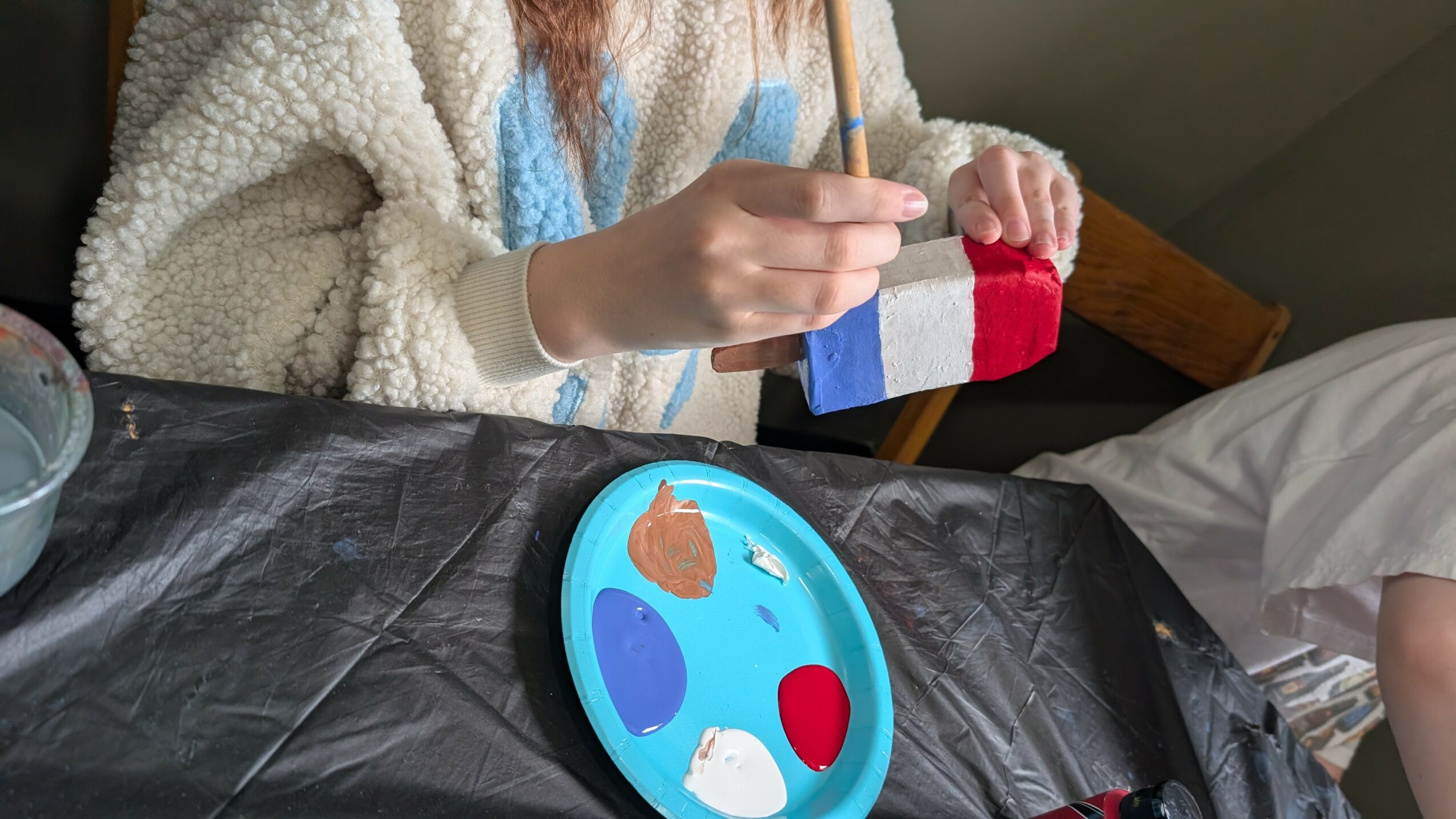 Person painting a small object with a paintbrush, sitting at a table with a blue palette, on a black tablecloth.