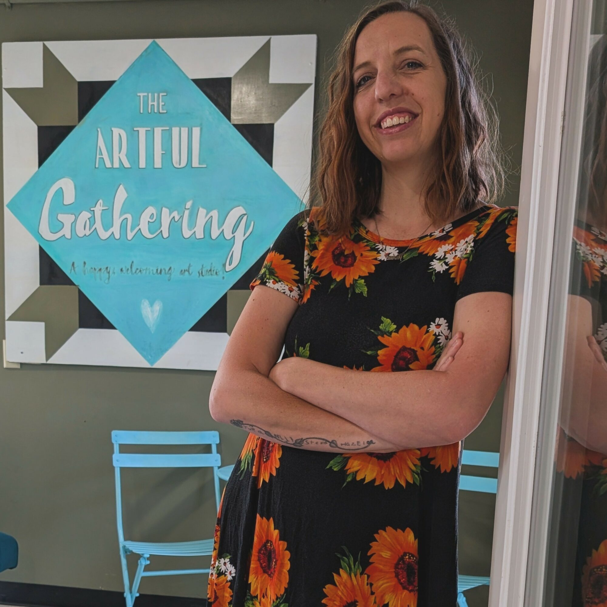 Woman with shoulder-length hair smiling, standing indoors near a window, wearing a black dress with orange and yellow floral pattern.