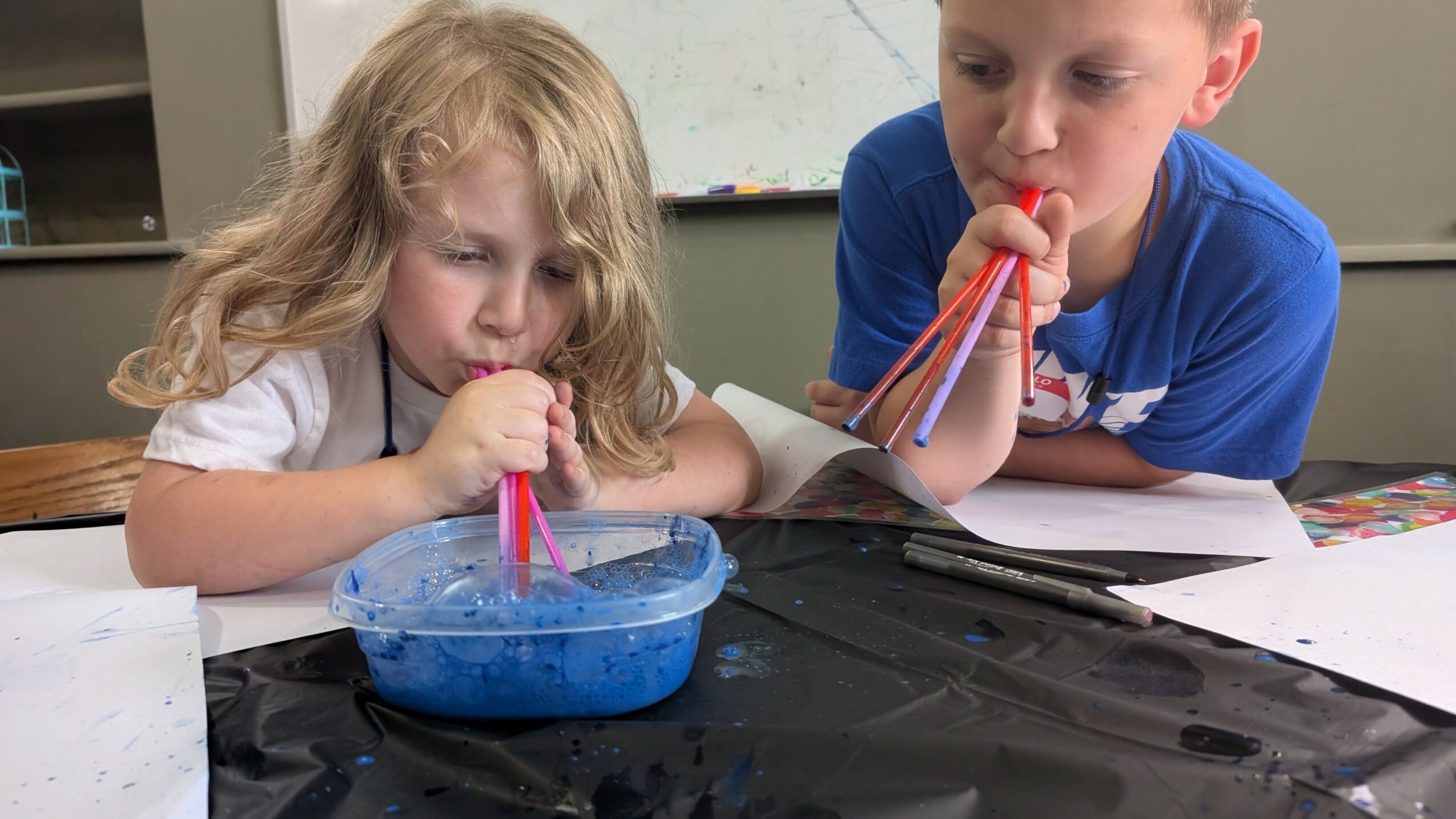 Two children blow bubbles into a blue container with straws, sitting at a table with papers and supplies.