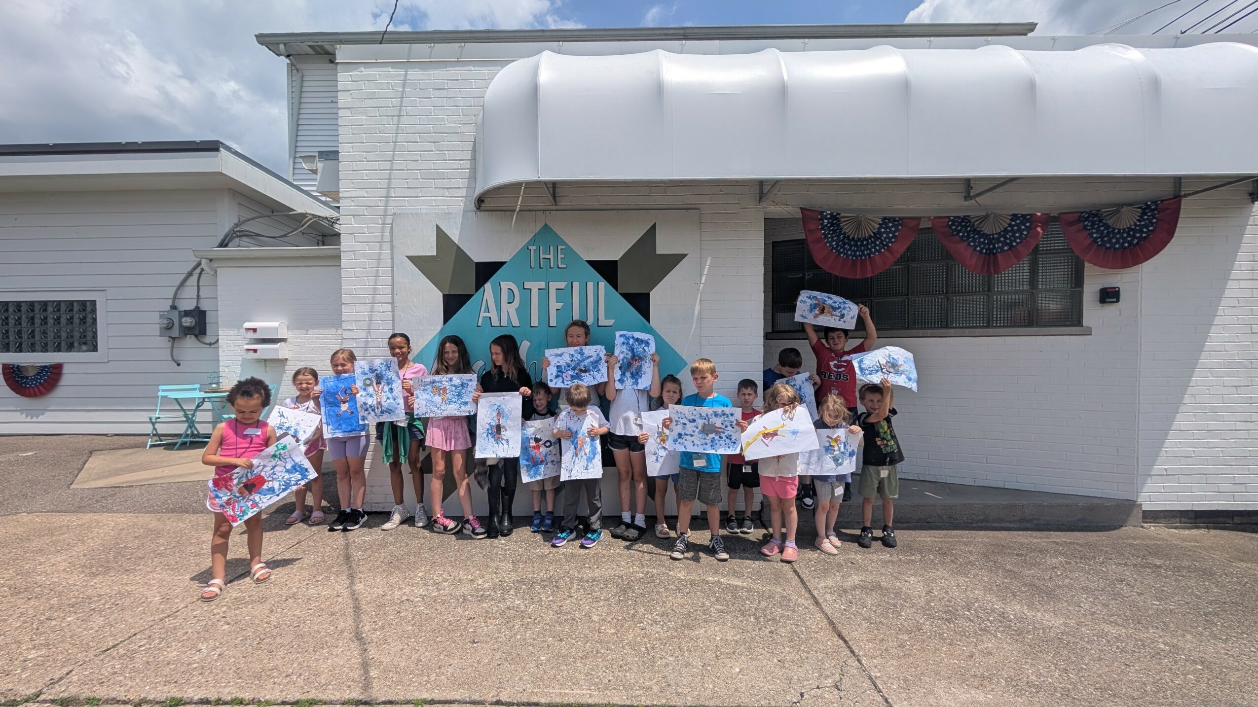 Group of children and adults standing outside a building holding signs and artwork, with a sign reading 'ARTFUL' behind them.