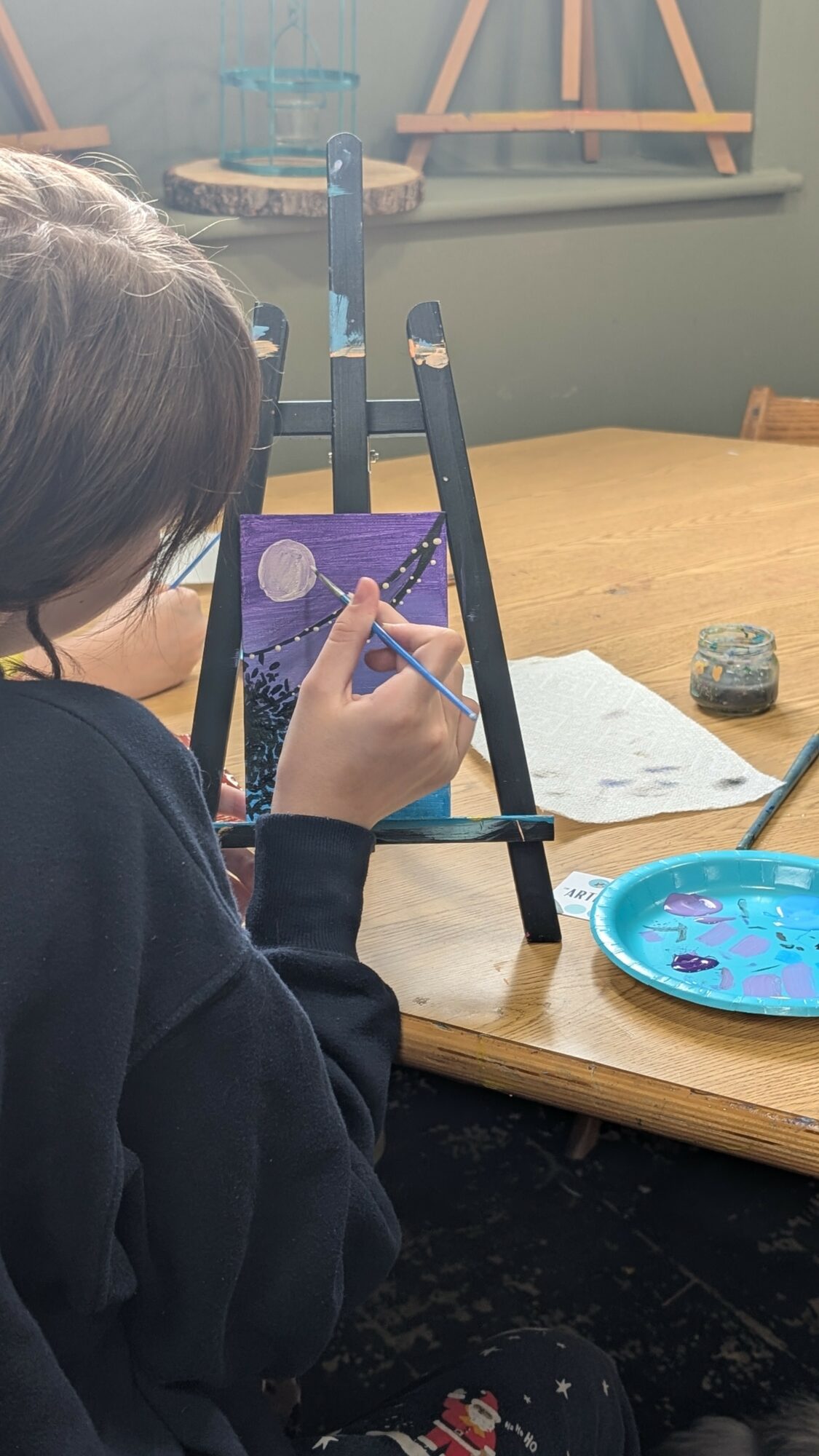 Child painting on small easel at table with art supplies, paper, and a blue palette, in a classroom setting.