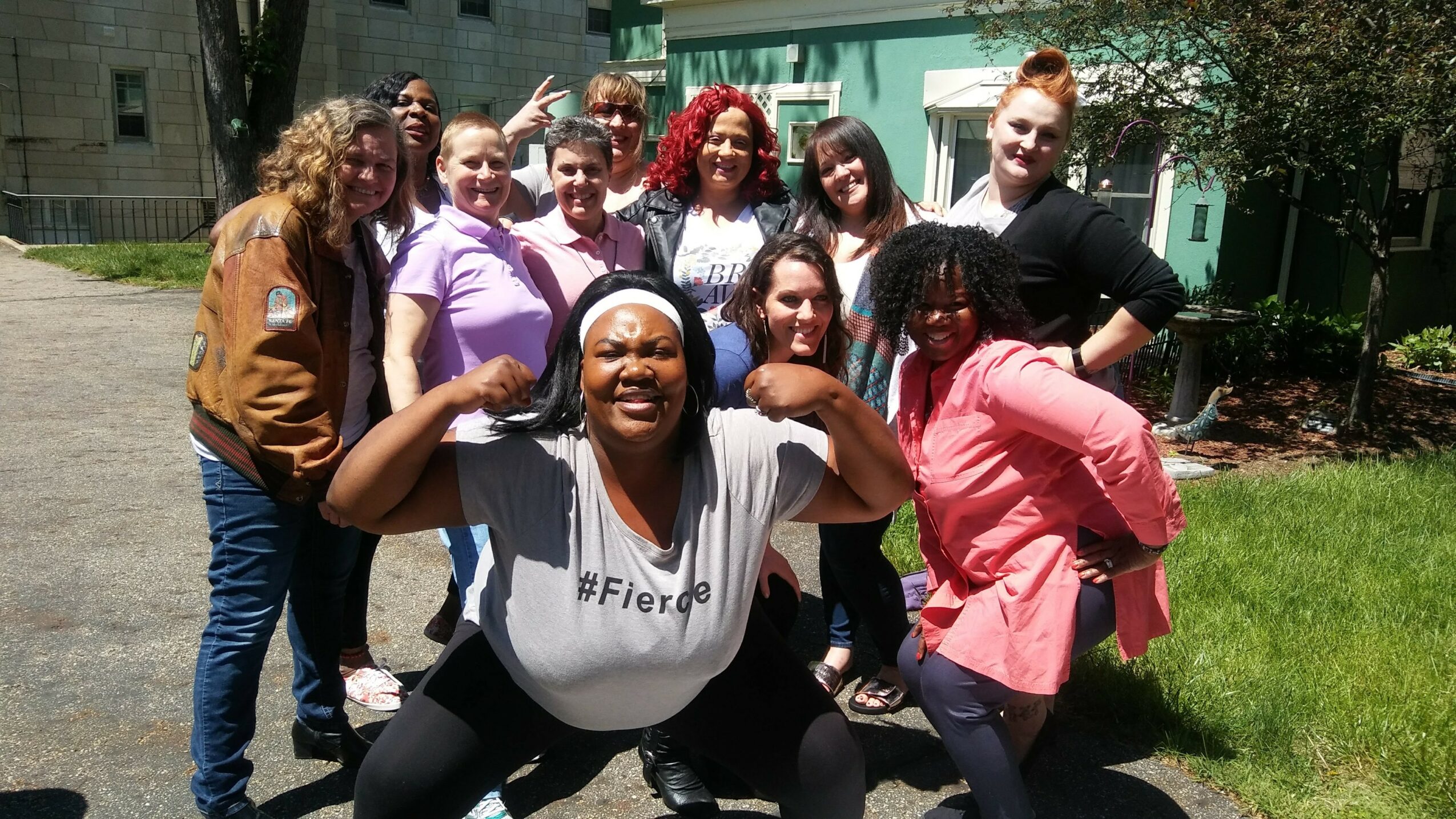 Group of diverse women smiling outdoors on a sunny day, some making gestures, in front of a building and greenery.