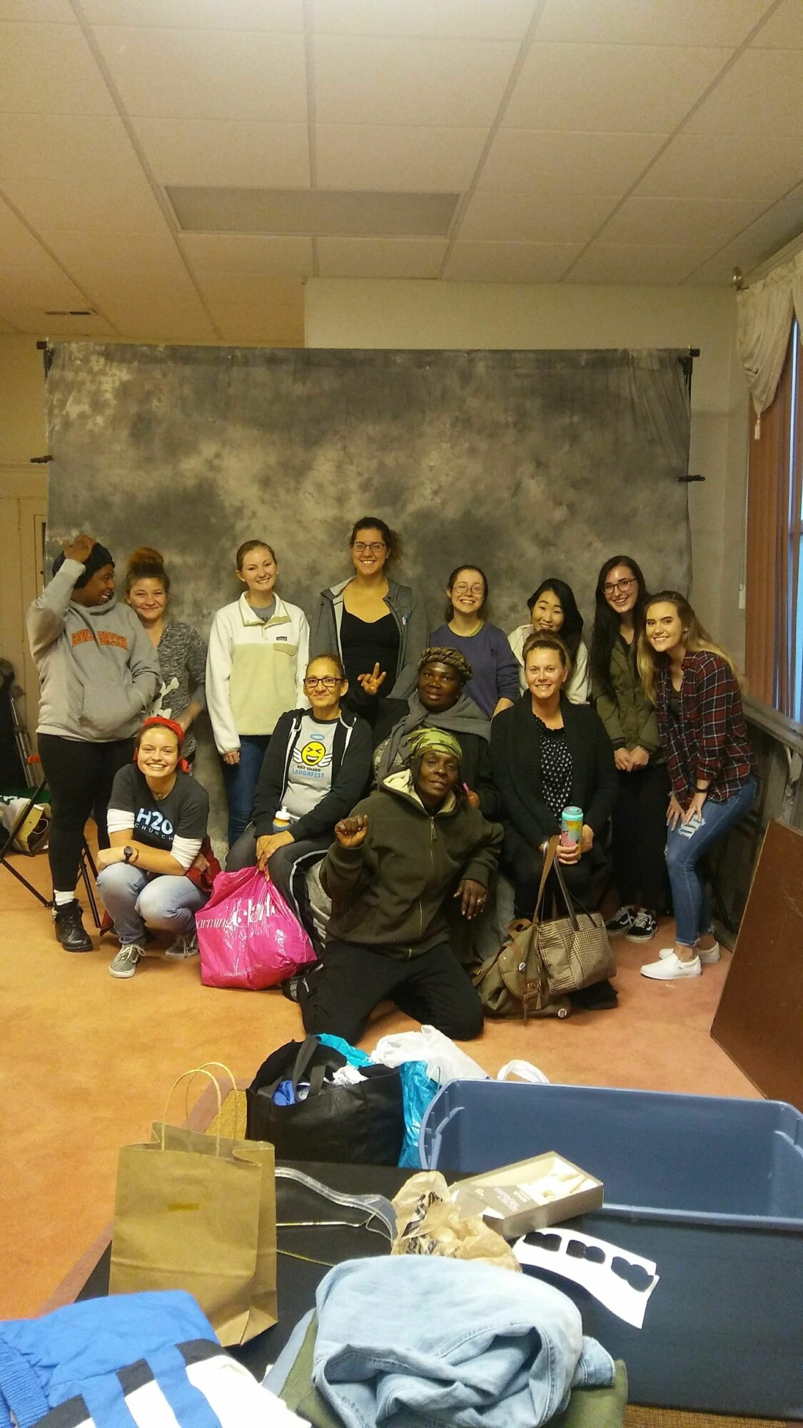 Group of people standing and sitting in a room with a gray wall and a blue table, some holding bags.