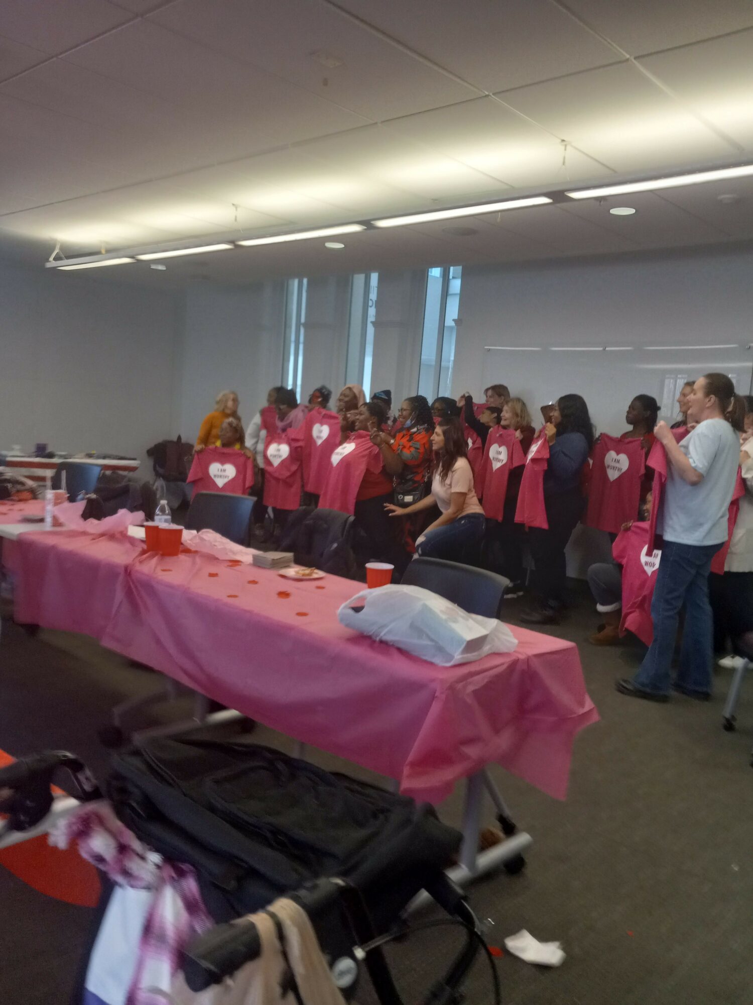 People gathered around a table with pink tablecloths in an indoor space, some standing and some sitting, with a bright ceiling and windows.