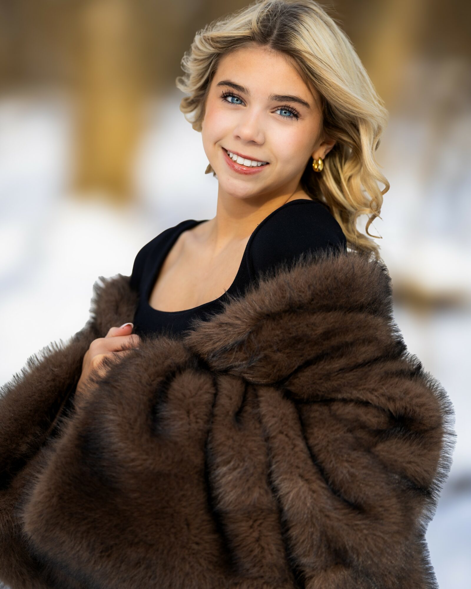 Young woman with blonde hair smiling outdoors in winter, wearing a black top and a brown fur coat.