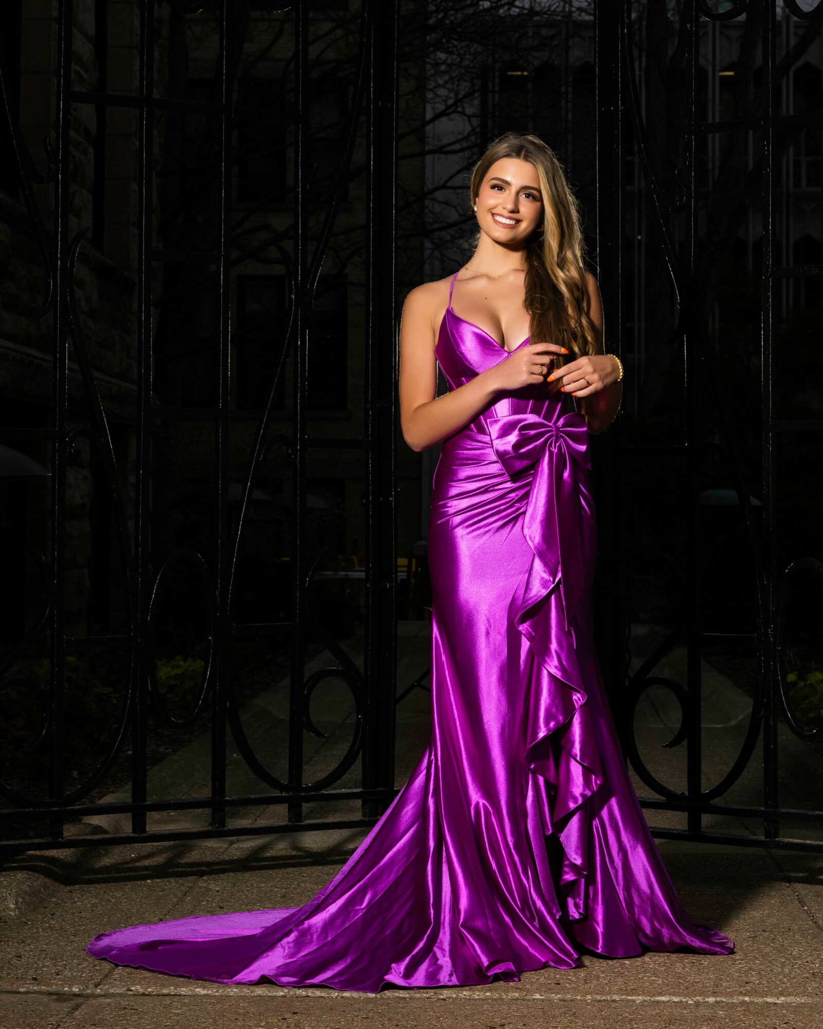 Young woman in a shiny purple gown standing in front of a black gate, smiling, with long wavy hair.