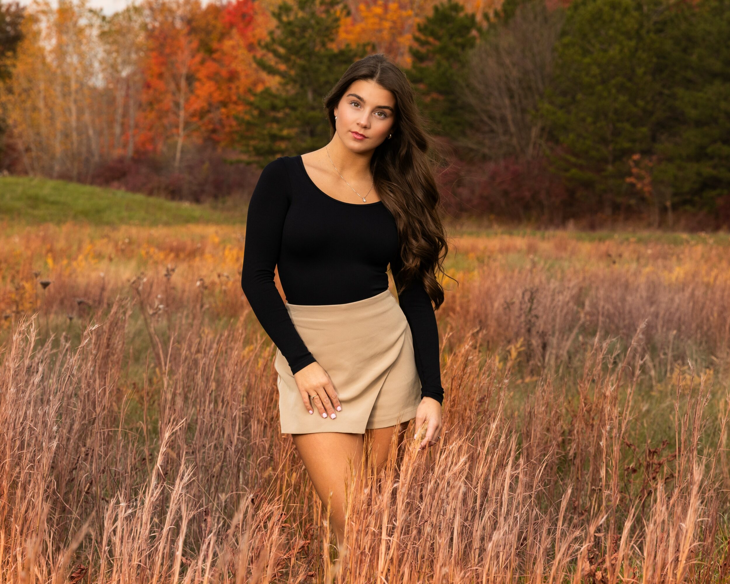 Young woman with long dark hair standing in a field with autumn trees in the background, wearing a black top and beige skirt.