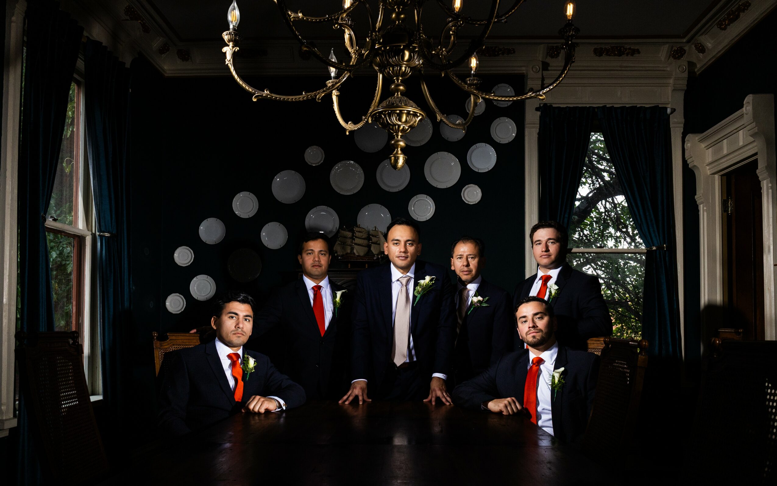 Six men in suits and ties pose in a dark, elegant room with a chandelier and large windows.