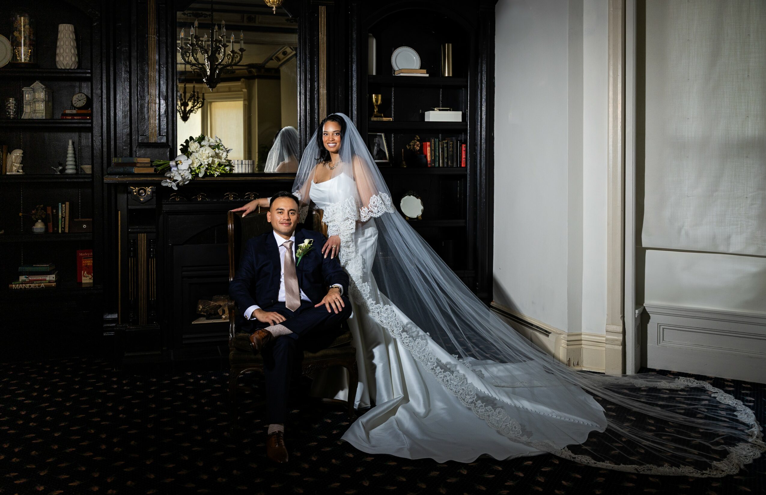 Bride in wedding dress and veil stands behind groom seated in a dark room with bookshelves, elegant decor, and a chandelier.