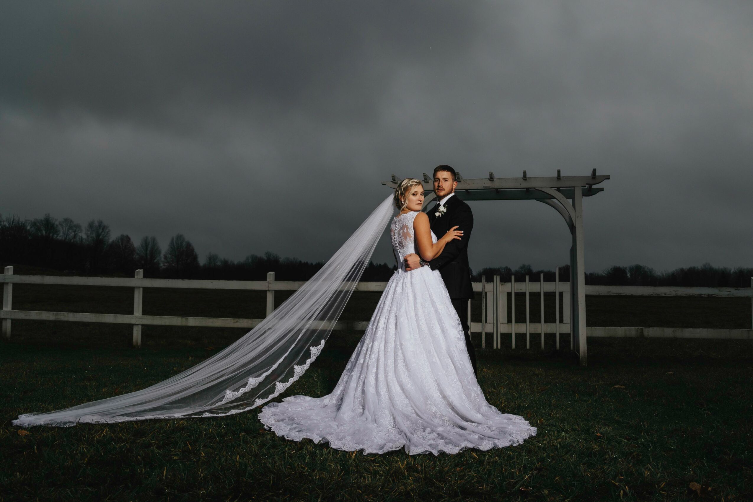 Bride and groom standing outdoors under dark stormy sky, bride in white gown with long veil, groom in black suit, fence in background.