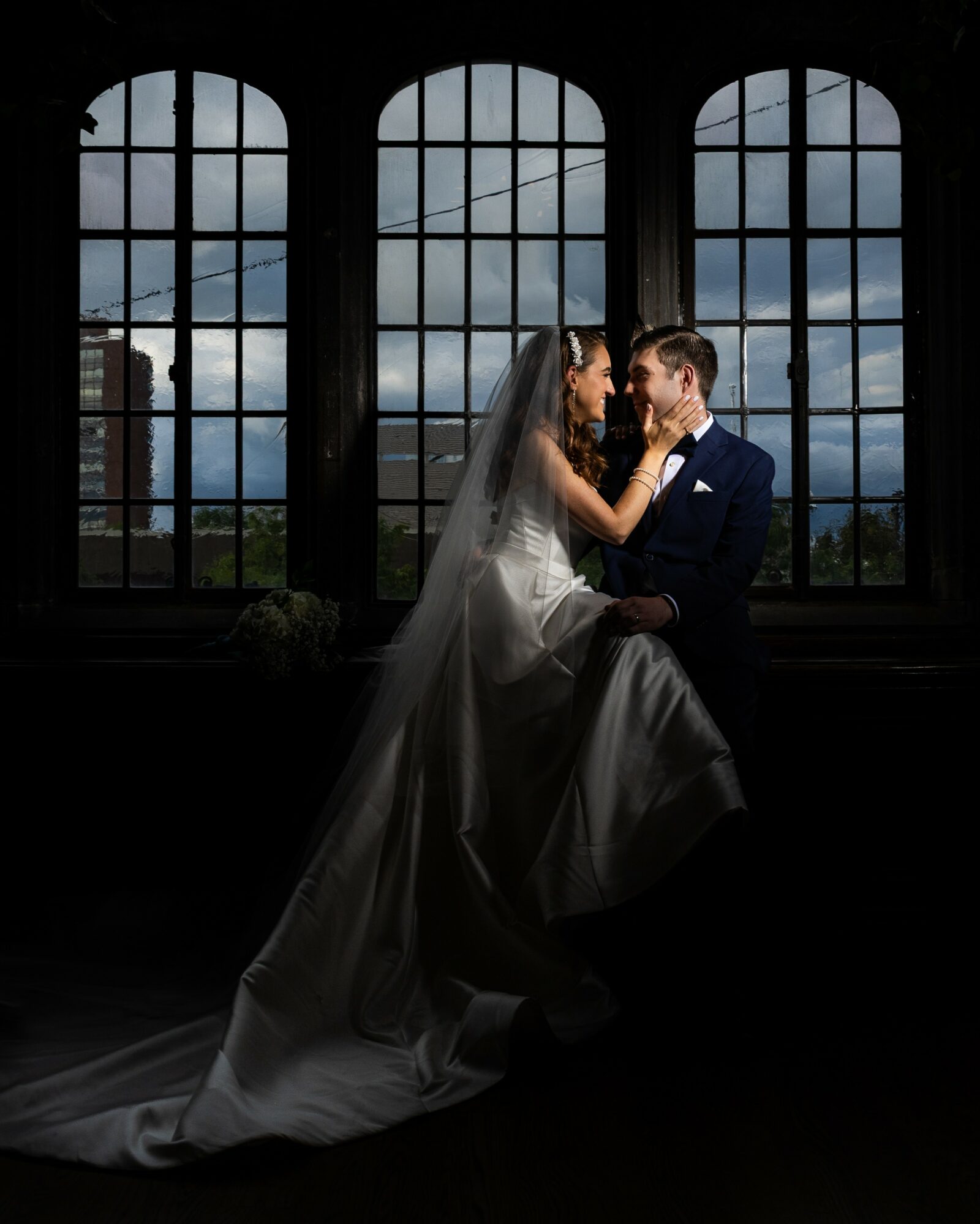 Bride and groom face each other indoors with large arched windows and cloudy sky background.