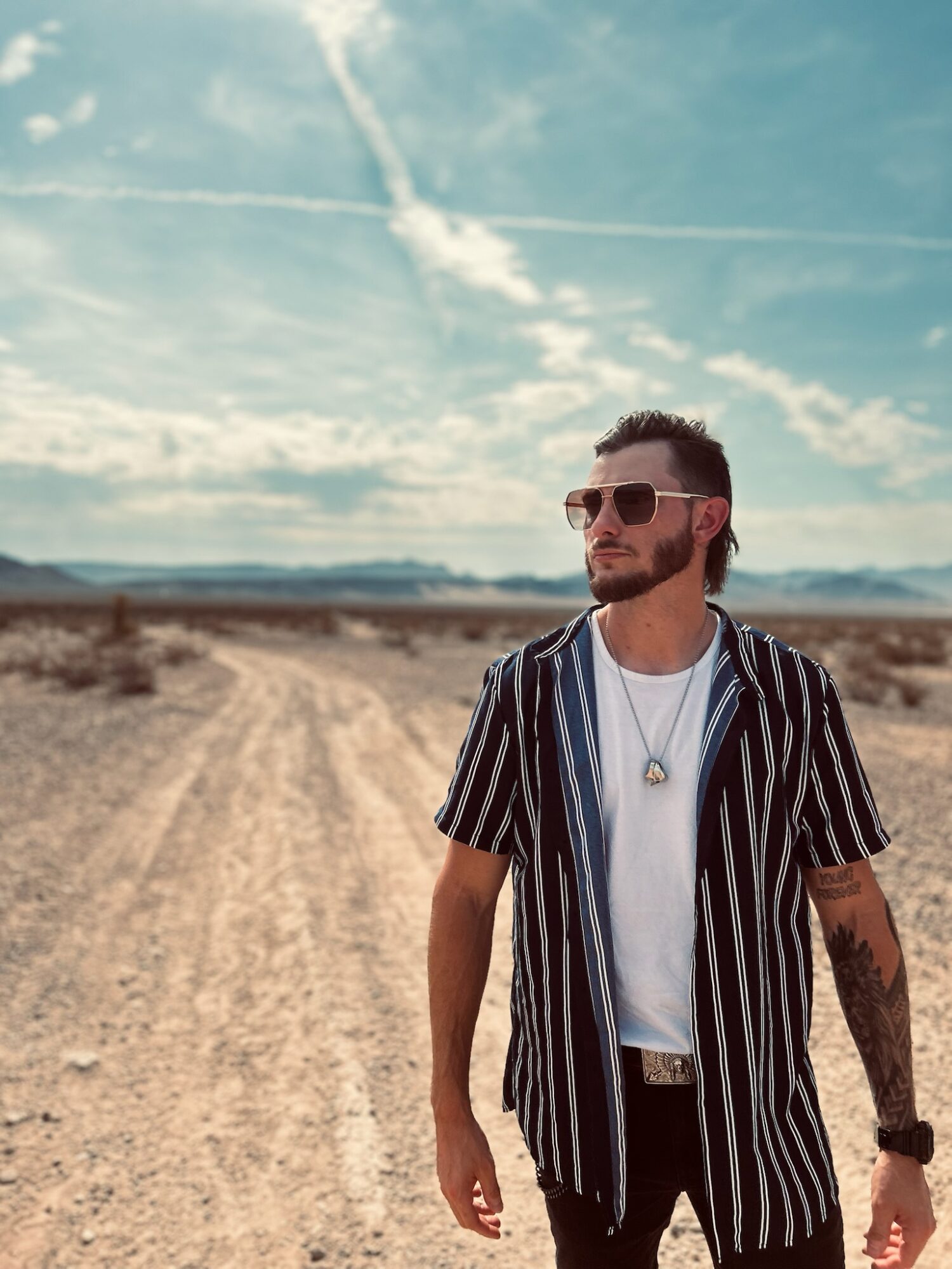 Man with sunglasses and striped shirt walking on a dirt road in a desert landscape under a blue sky with clouds.