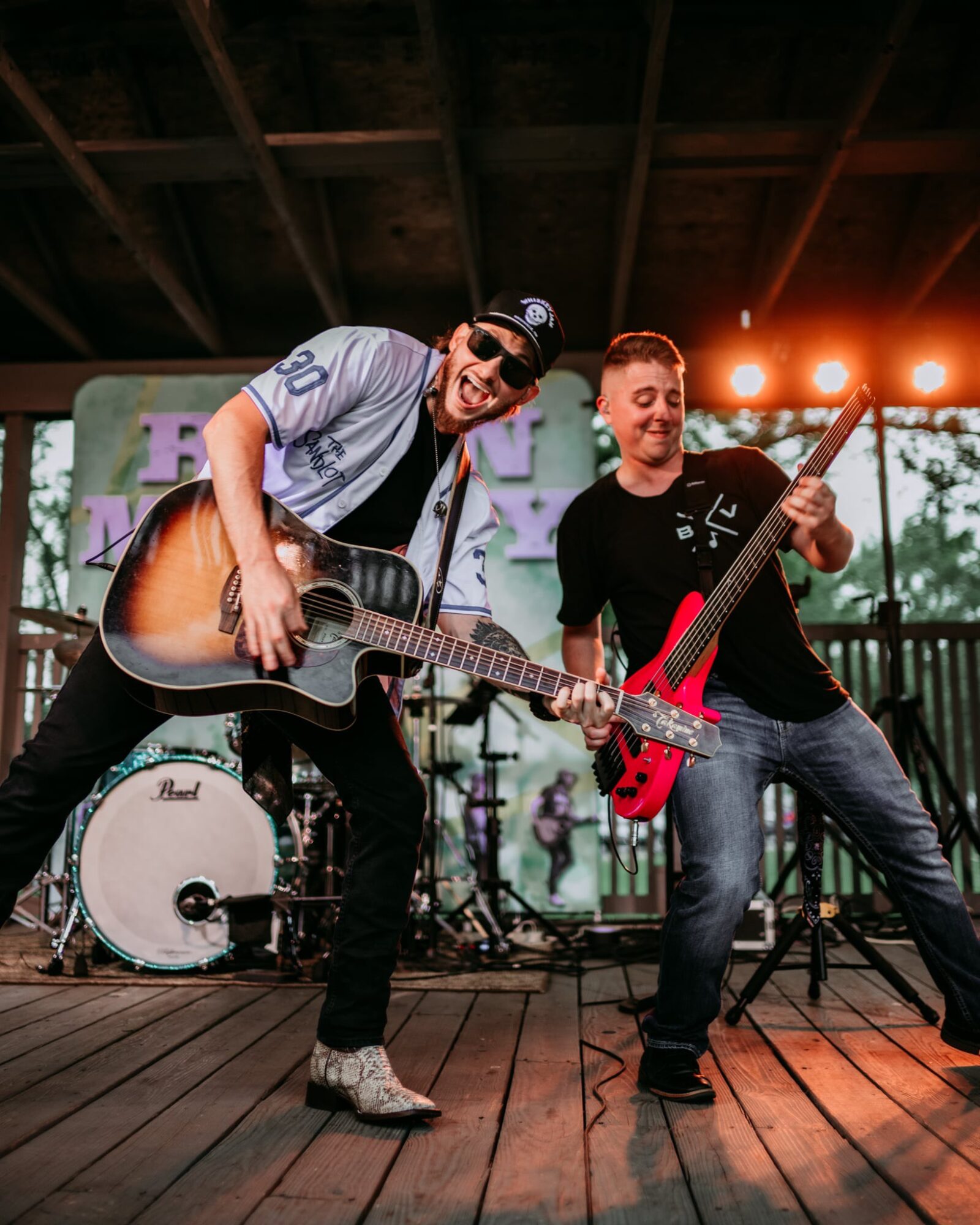 Two musicians perform on stage with guitars, smiling, under stage lights, with a drum set in the background.