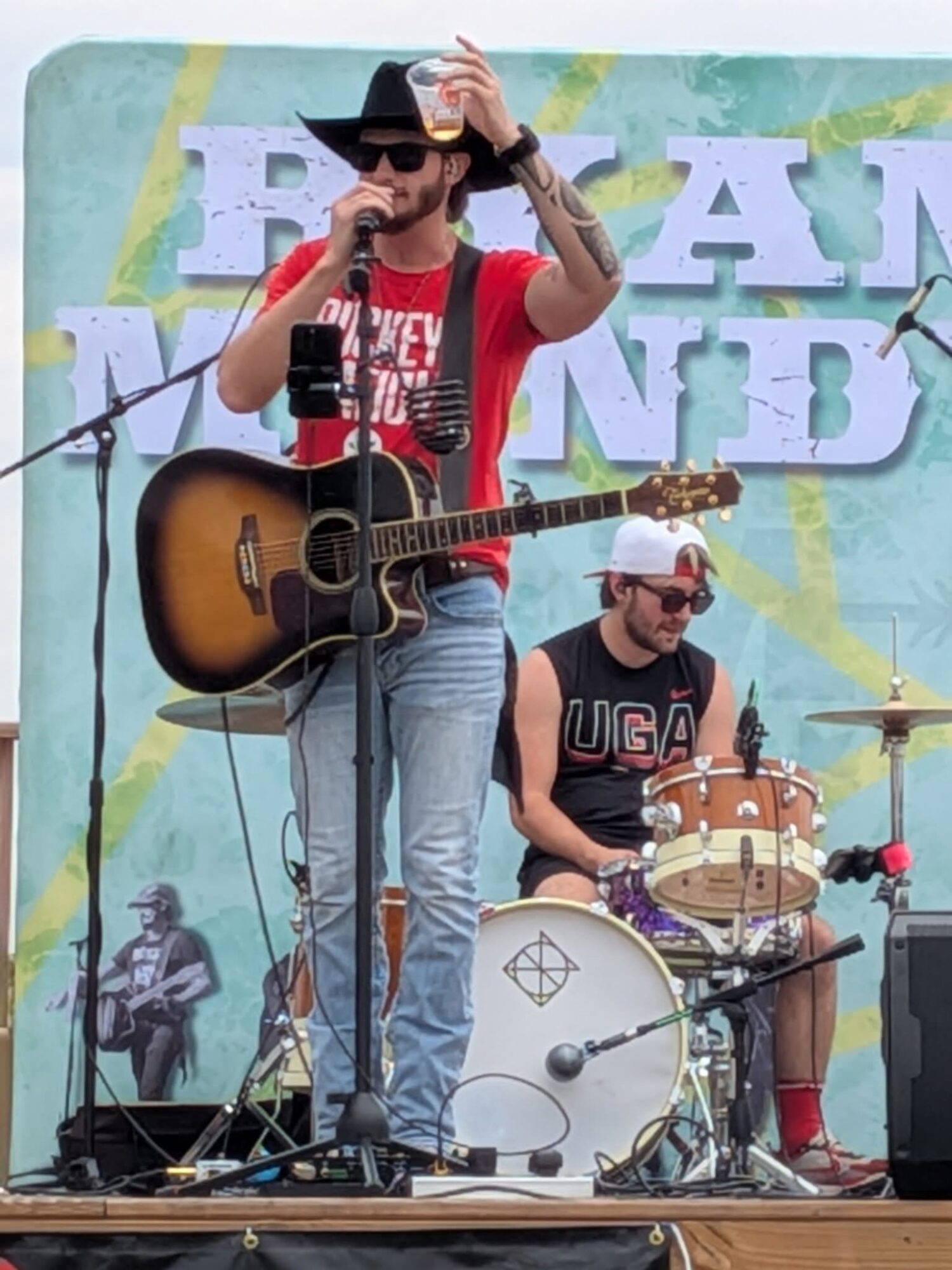 Two musicians perform on stage, one singing with a guitar, the other playing drums, with a colorful backdrop.