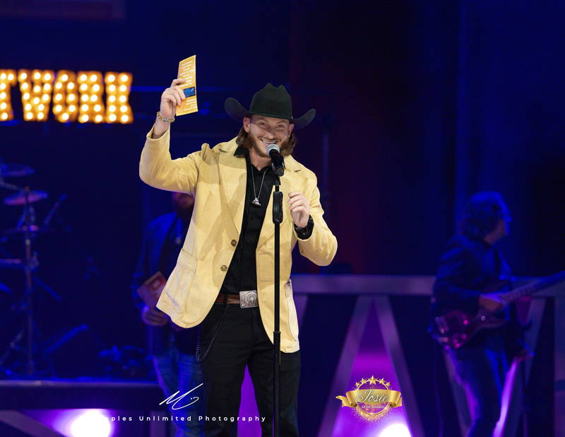 Person in yellow jacket and cowboy hat singing on stage with a microphone, background dark with blue lighting.