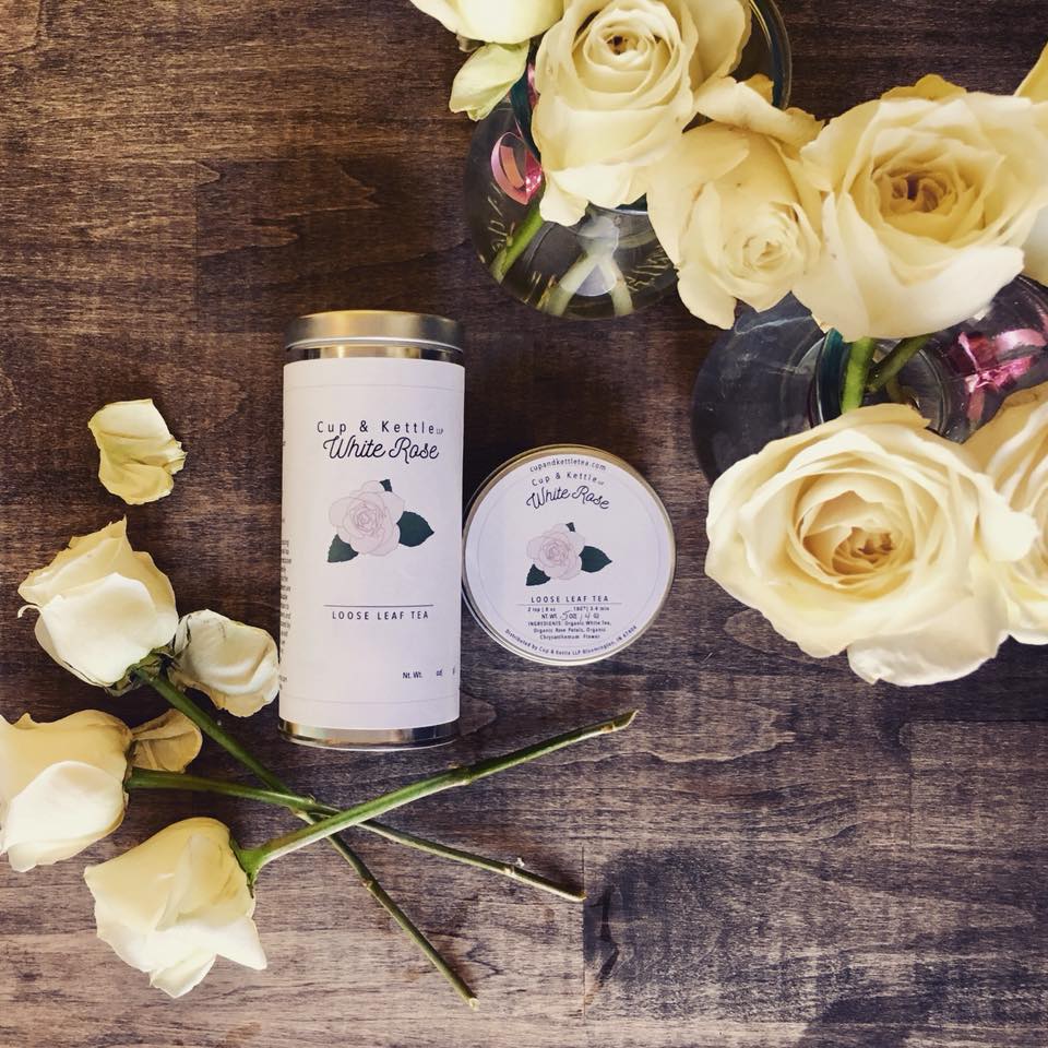 White roses and a candle on a wooden surface, with a small jar and a tin container nearby.