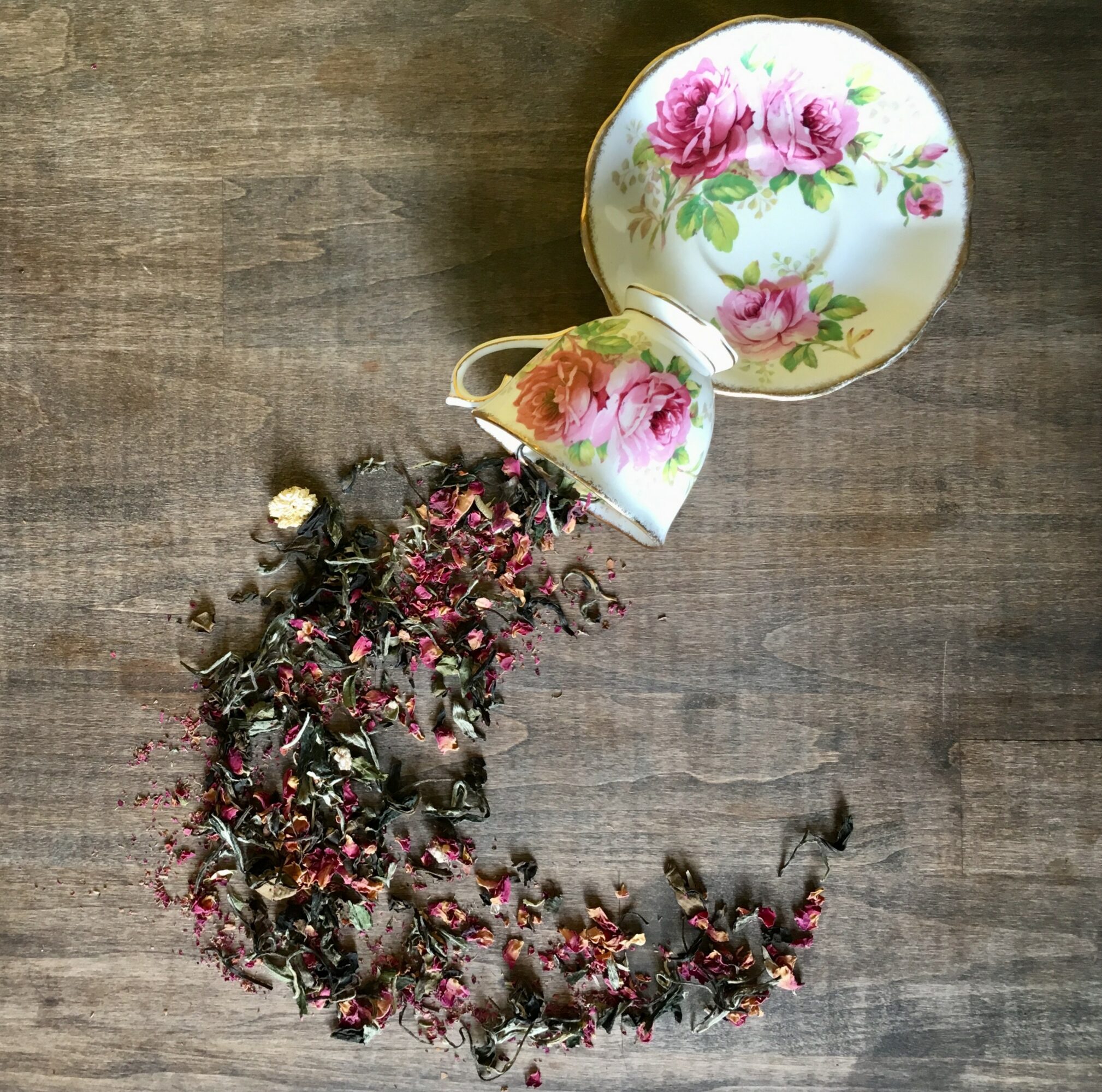 Broken floral teacup and saucer with pink roses on a wooden surface, with dried flower petals and debris.