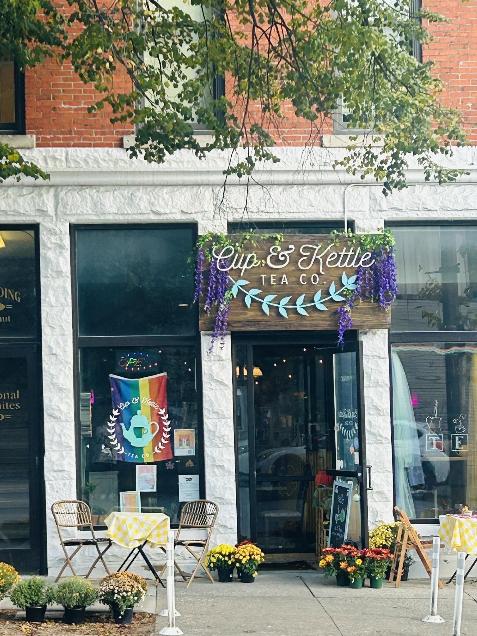 Storefront with sign 'Cup & Kettle Tea Co.', purple flowers, outdoor seating, and potted plants outside.