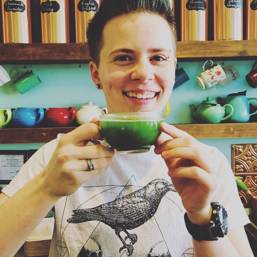 Person smiling holding a green cup in a colorful tea shop with teapots on shelves behind.