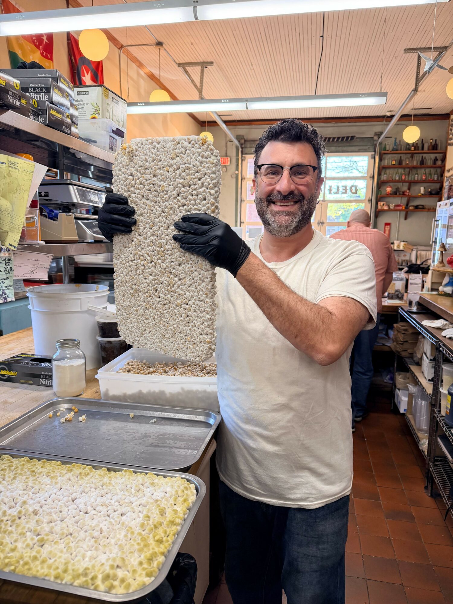 Man with dark hair and beard, wearing white t-shirt and black gloves, stands next to a large block of rice, in a kitchen or food preparation area.