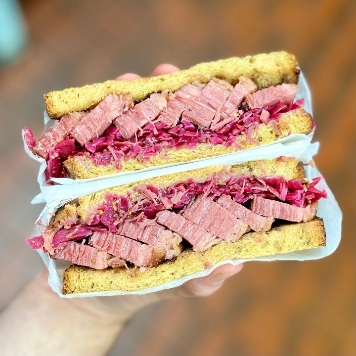 Person holding a large sandwich with sliced meat and shredded vegetables between bread slices, wrapped in paper, against a blurred background.