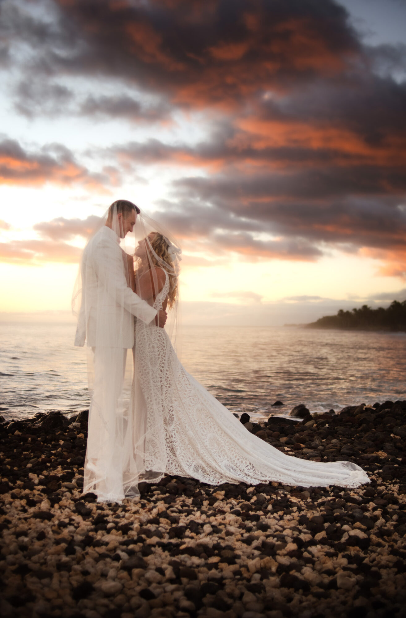 A couple in wedding attire stands on a rocky beach at sunset, holding hands and facing each other.